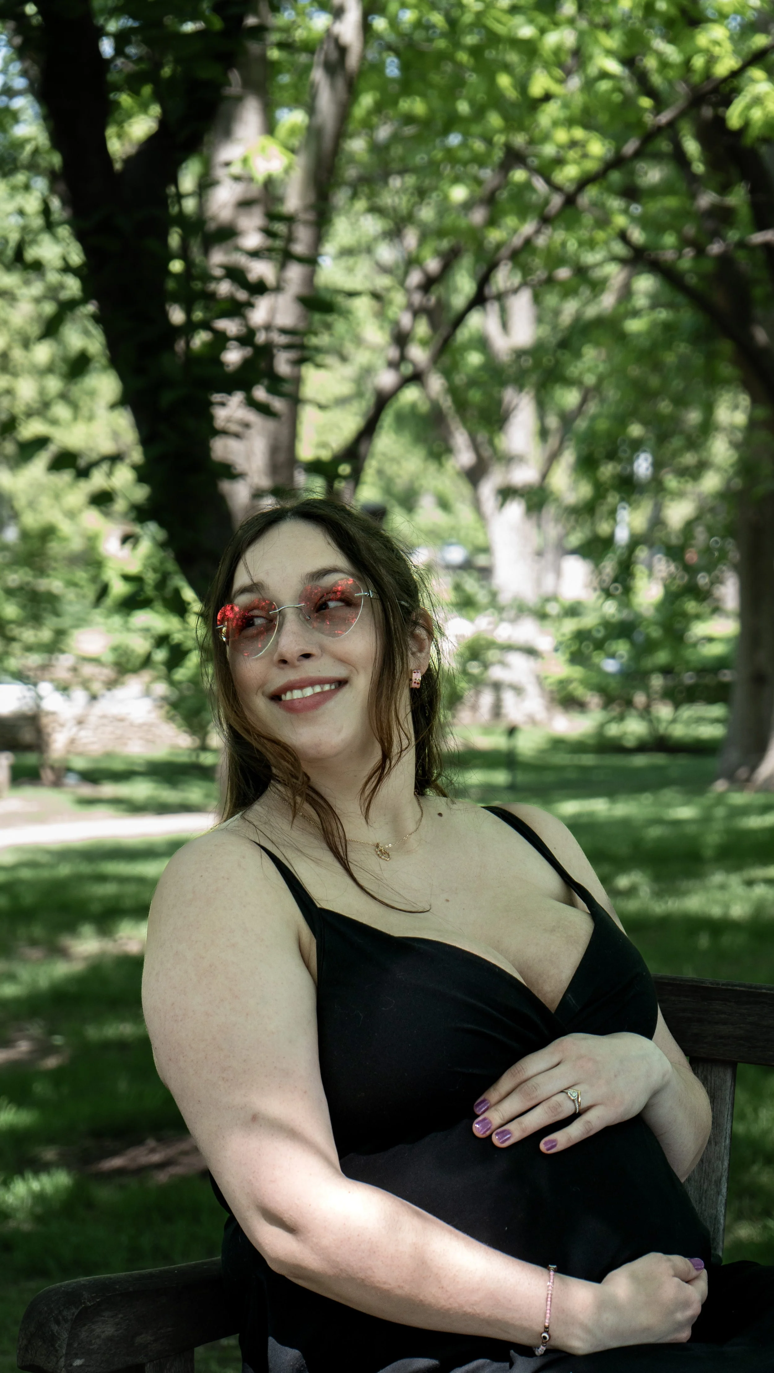 A smiling woman with wavy brown hair and glasses sitting outdoors on a bench in a park with green trees in the background, wearing a black sleeveless top and jewelry.