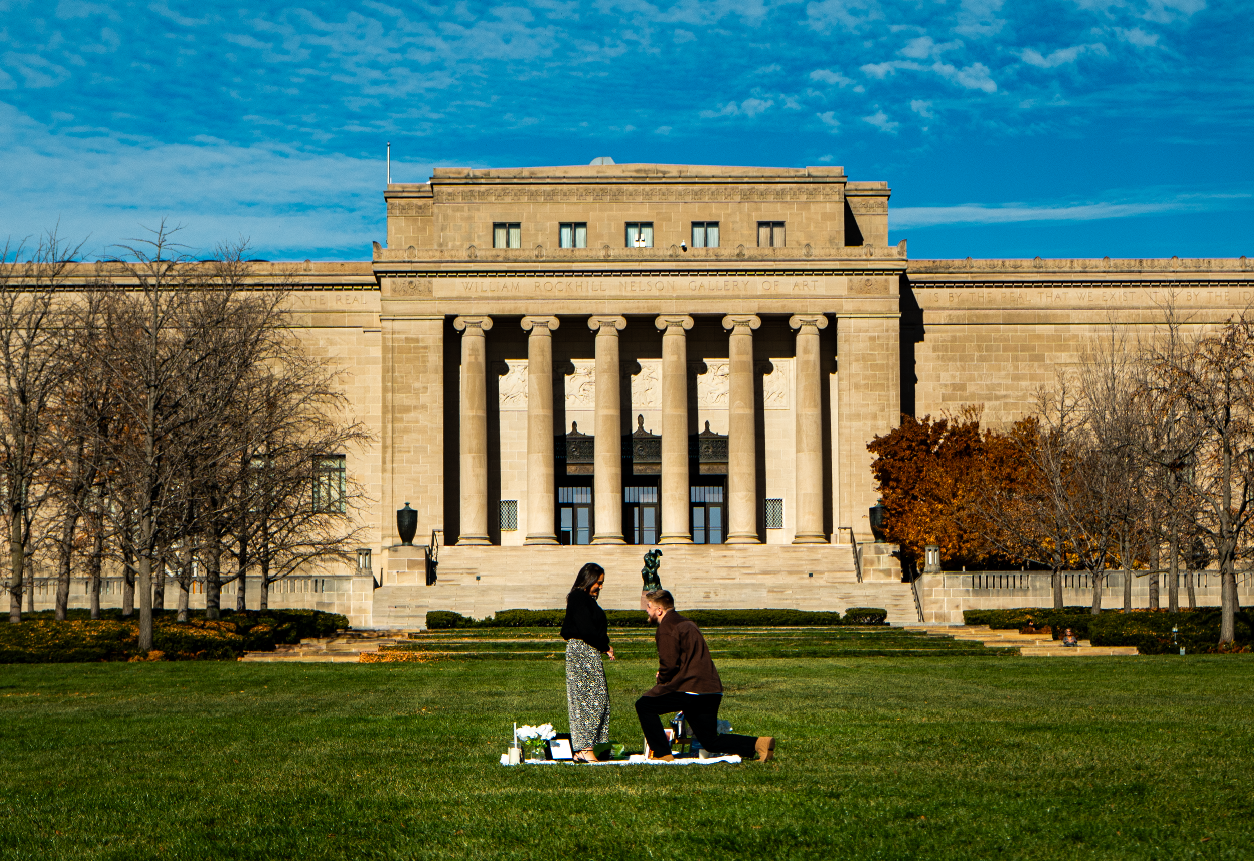 A couple participating in a wedding ceremony outdoors in front of the William Rockhill Nelson Gallery of Art building, with trees and a blue sky in the background.