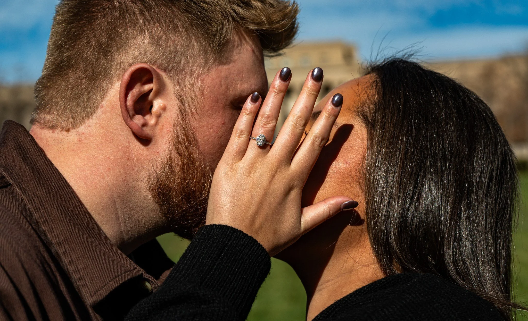 A close-up of a man and woman sharing a kiss outdoors, with the woman's hand showing a diamond engagement ring.