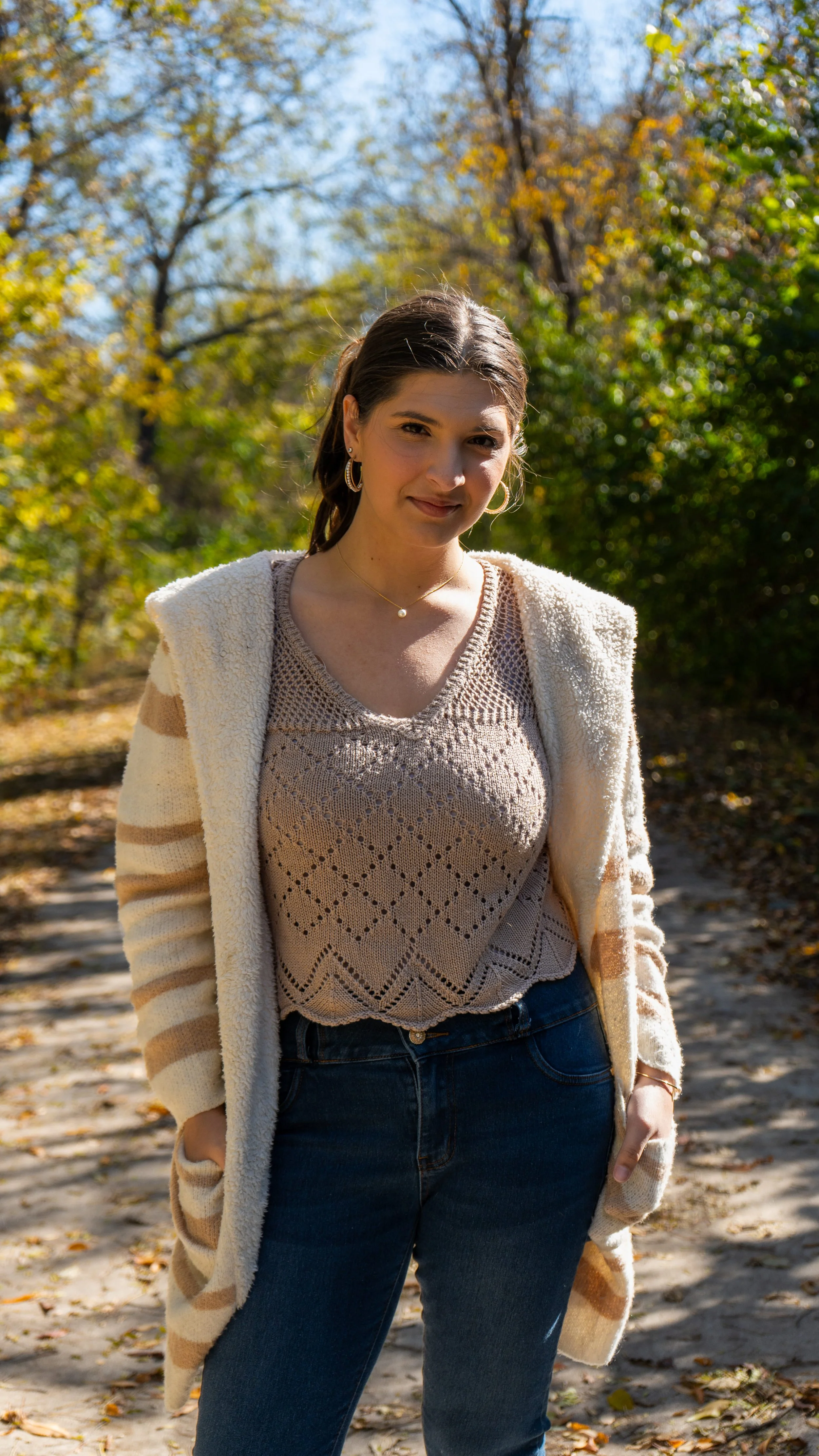 A woman standing outdoors in a park on a sunny autumn day, wearing a beige patterned top, dark jeans, and a cream-colored cardigan, with trees and fallen leaves in the background.