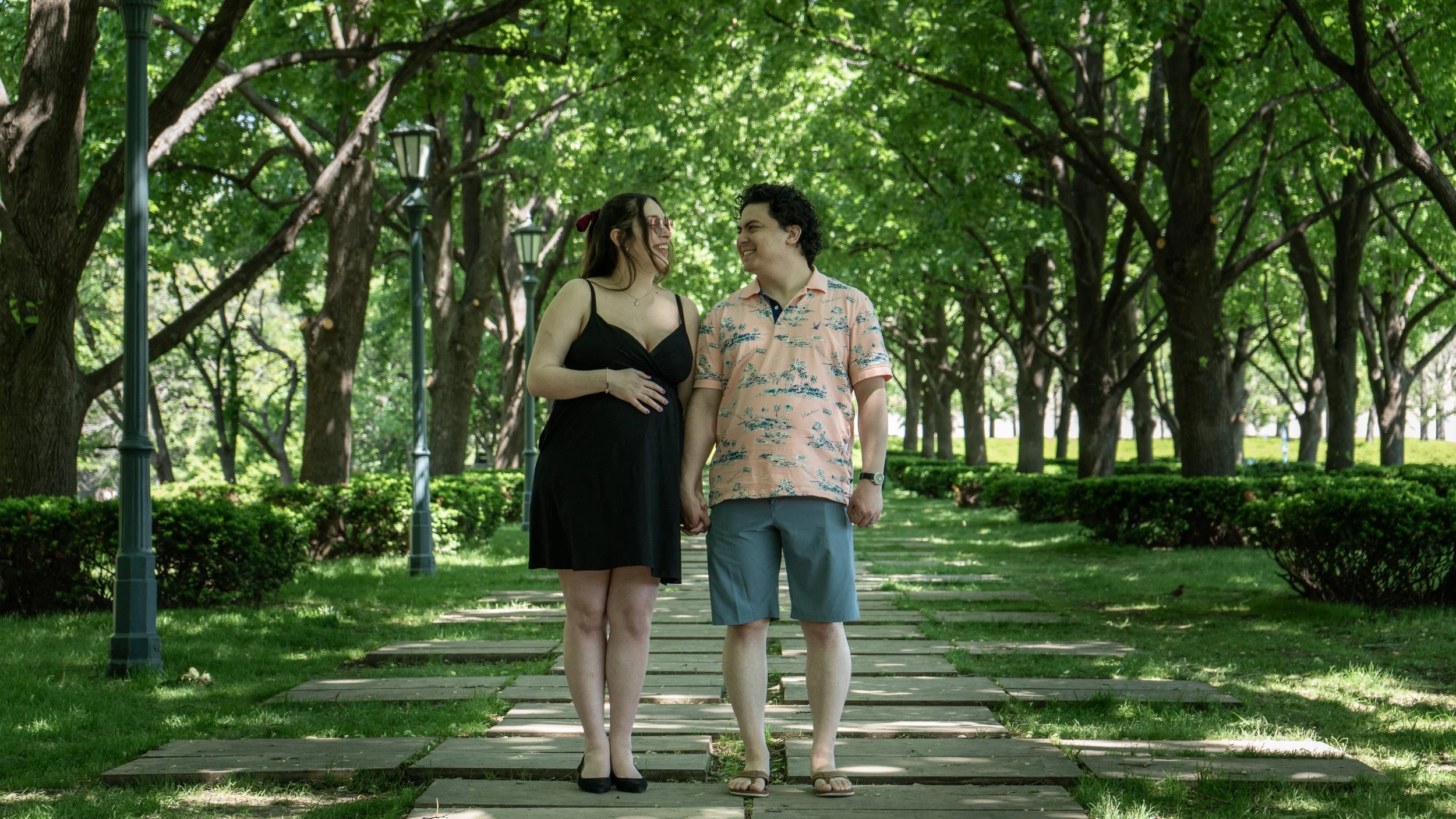 A pregnant woman and a man holding hands walk together on a tree-lined path in a park, smiling at each other.