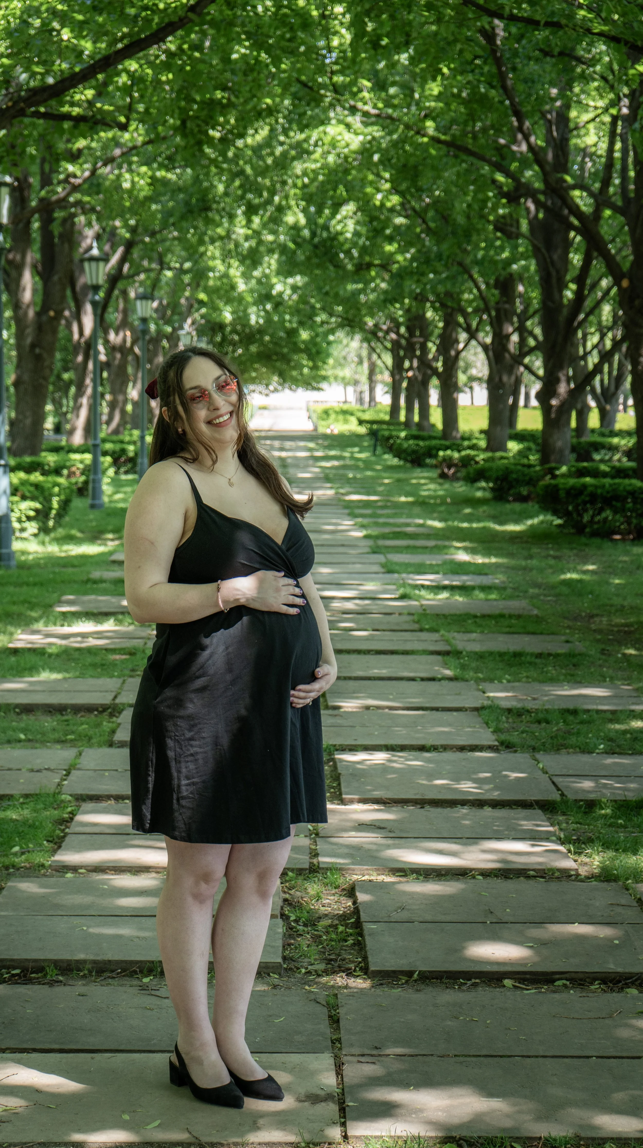 A pregnant woman stands outside on a stone pathway lined with trees and bushes, holding her belly and smiling.