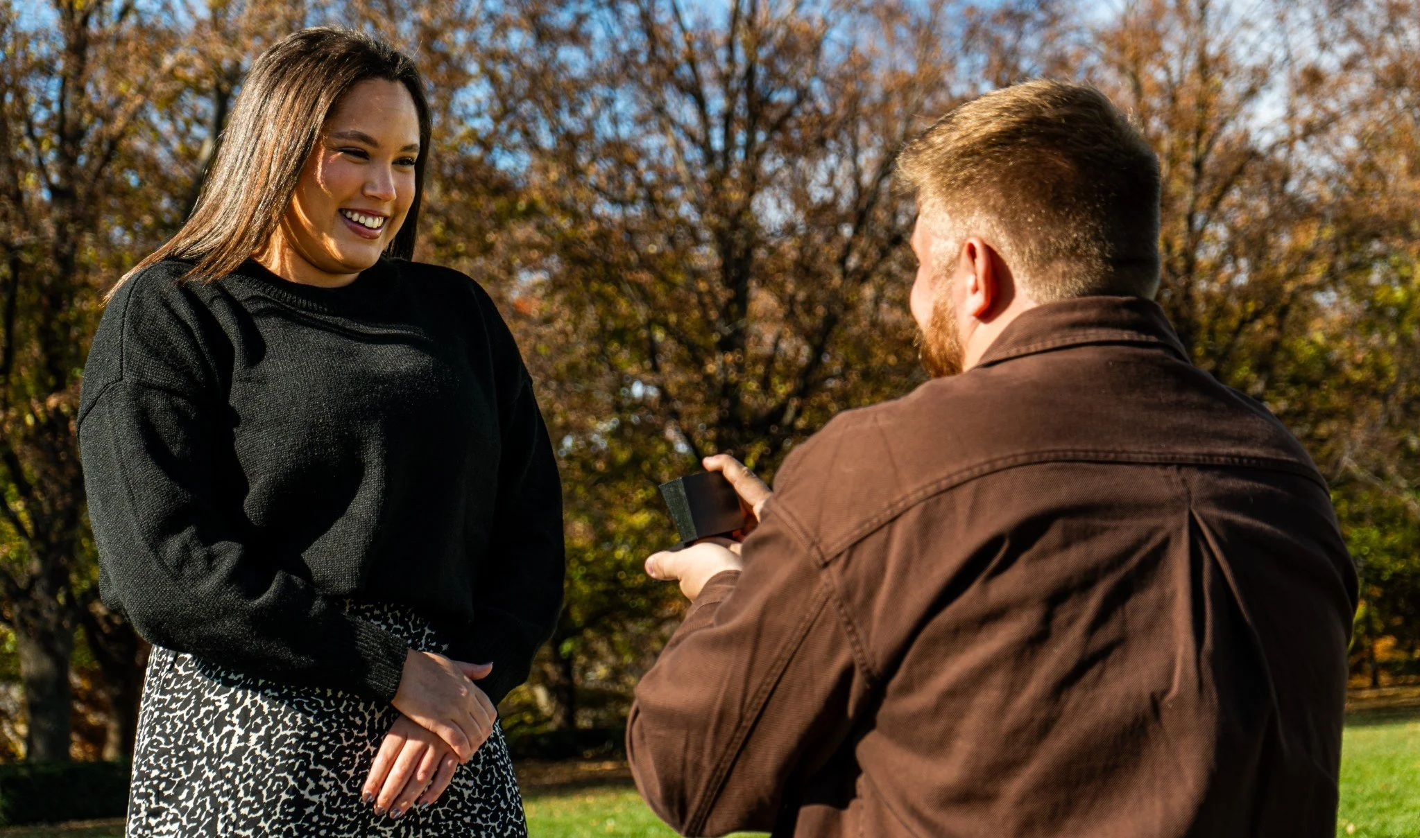 A woman with long brown hair smiling and looking at a man who is holding a small black box, with a background of trees with fall-colored leaves.