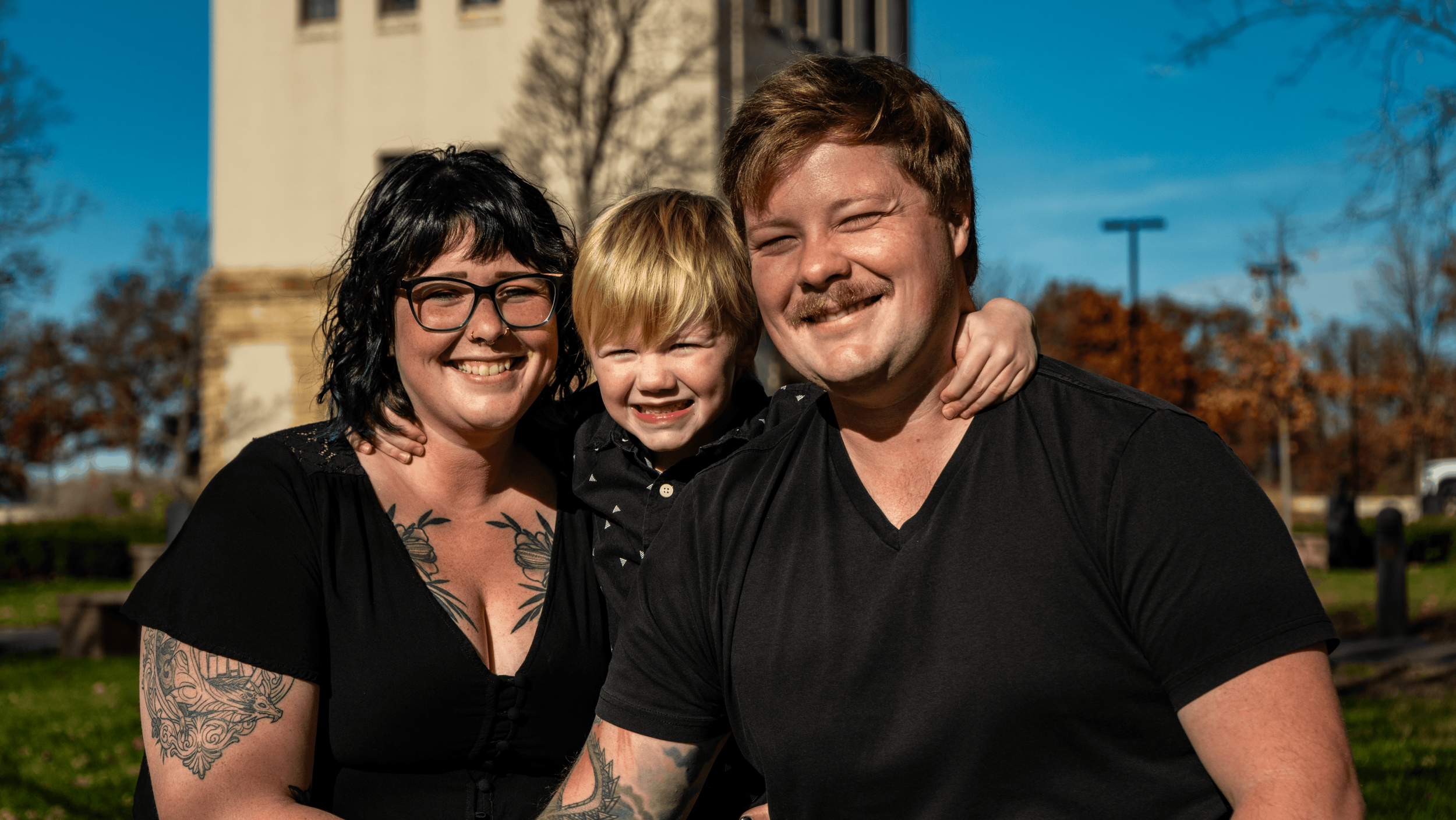 A group of three smiling people, two adults and a child, standing outdoors with a building and trees with fall foliage in the background.