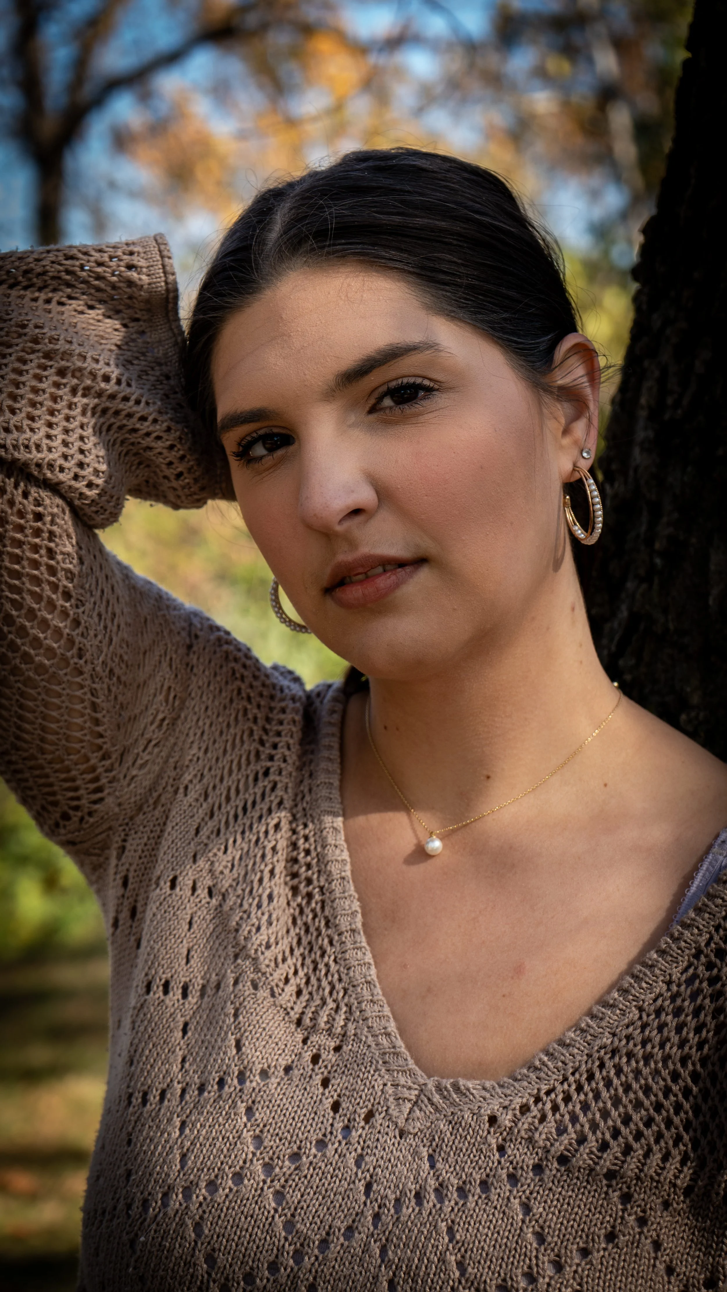 A young woman with dark hair and light skin poses outdoors near a tree, wearing a beige, open-knit sweater, pearl necklace, and hoop earrings, with autumn foliage in the background.