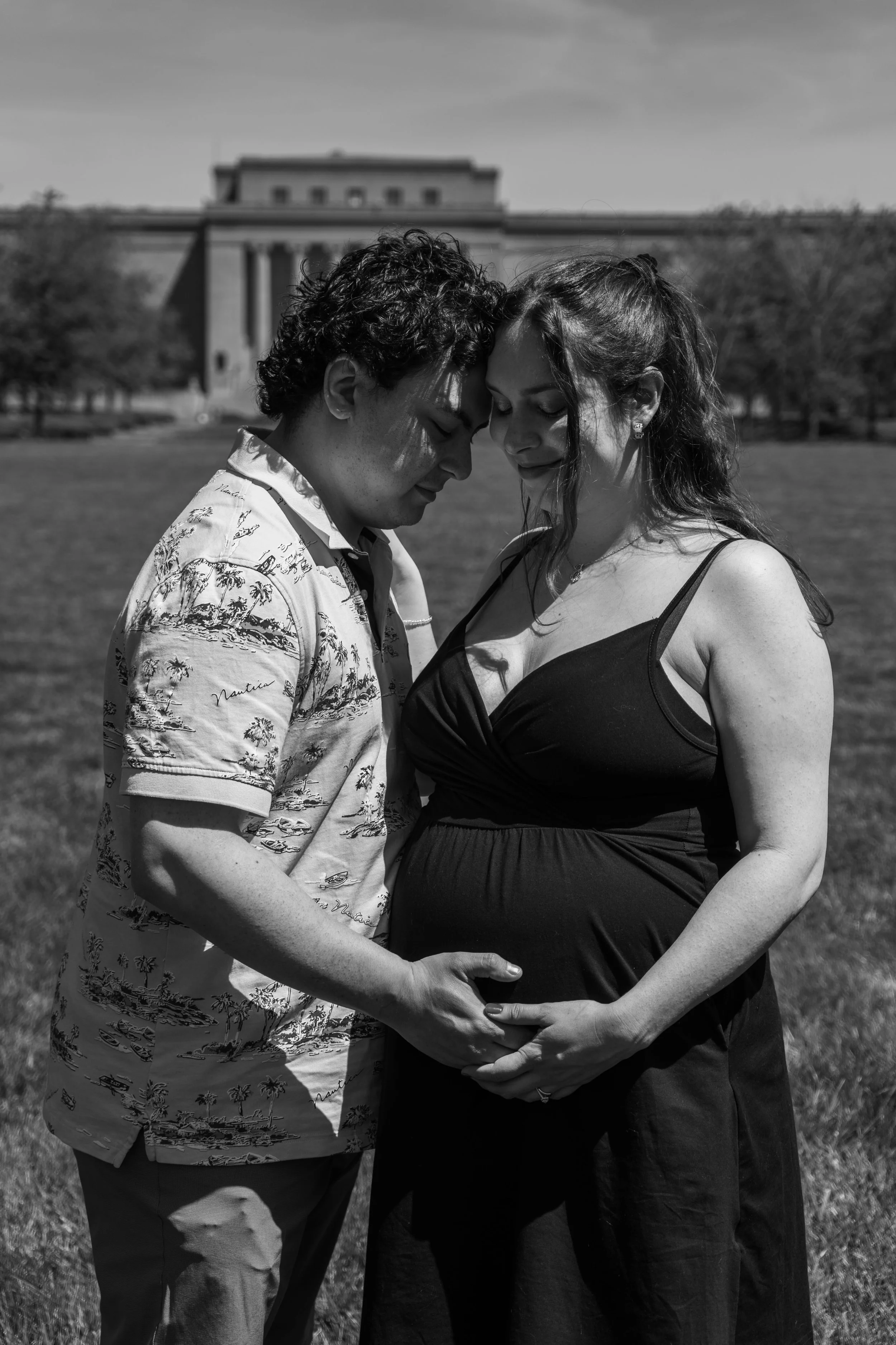 Black and white photo of a pregnant woman and a man standing outdoors with trees and a large building in the background. They are touching foreheads and holding her baby bump, with their eyes closed and gentle expressions.