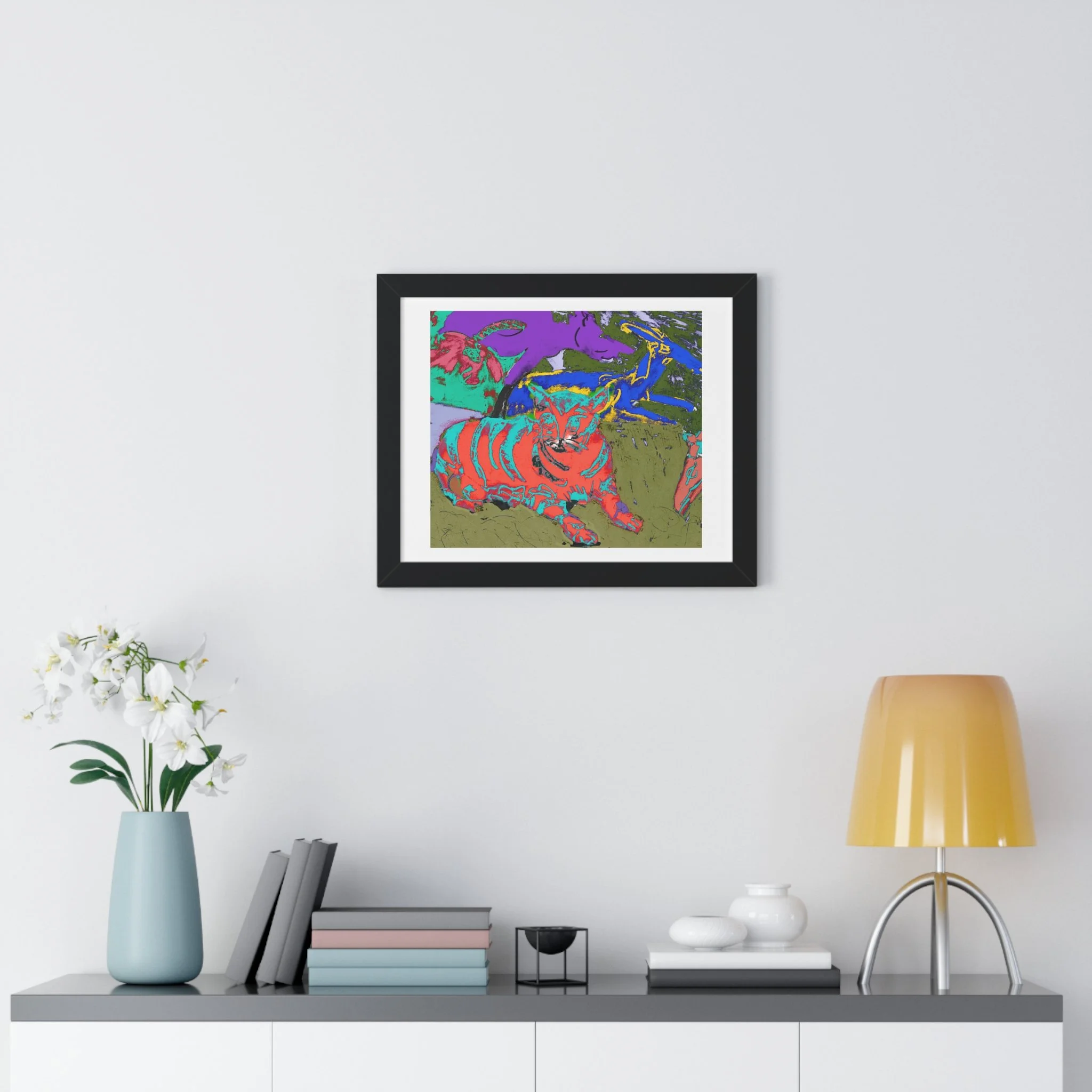 Colorful cat art print framed in black with a matte.  hang above a table with light blue vase and white flower, books, and lamp.