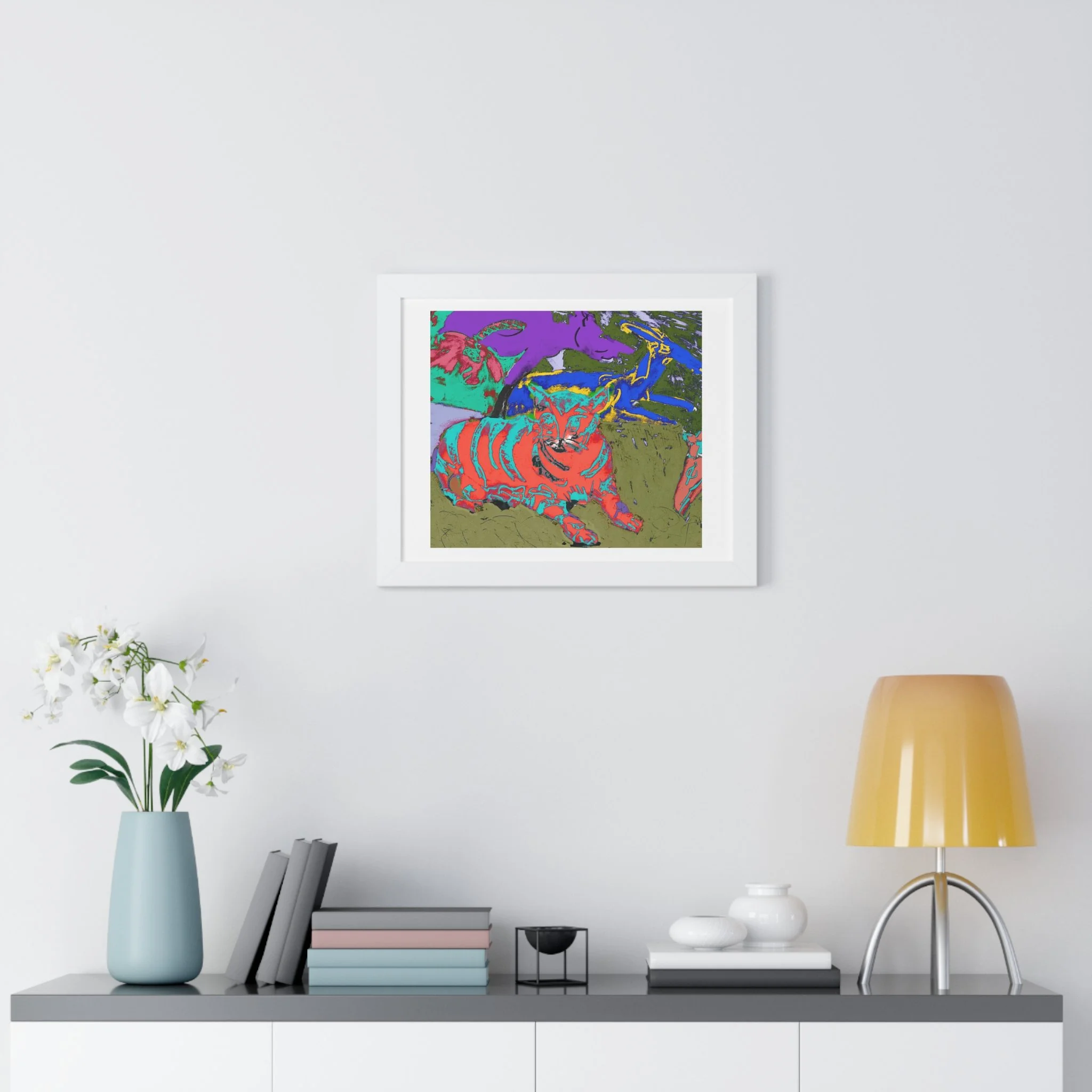 Colorful cat art print framed in white with a matte.  hang above a table with light blue vase and white flower, books, and lamp.