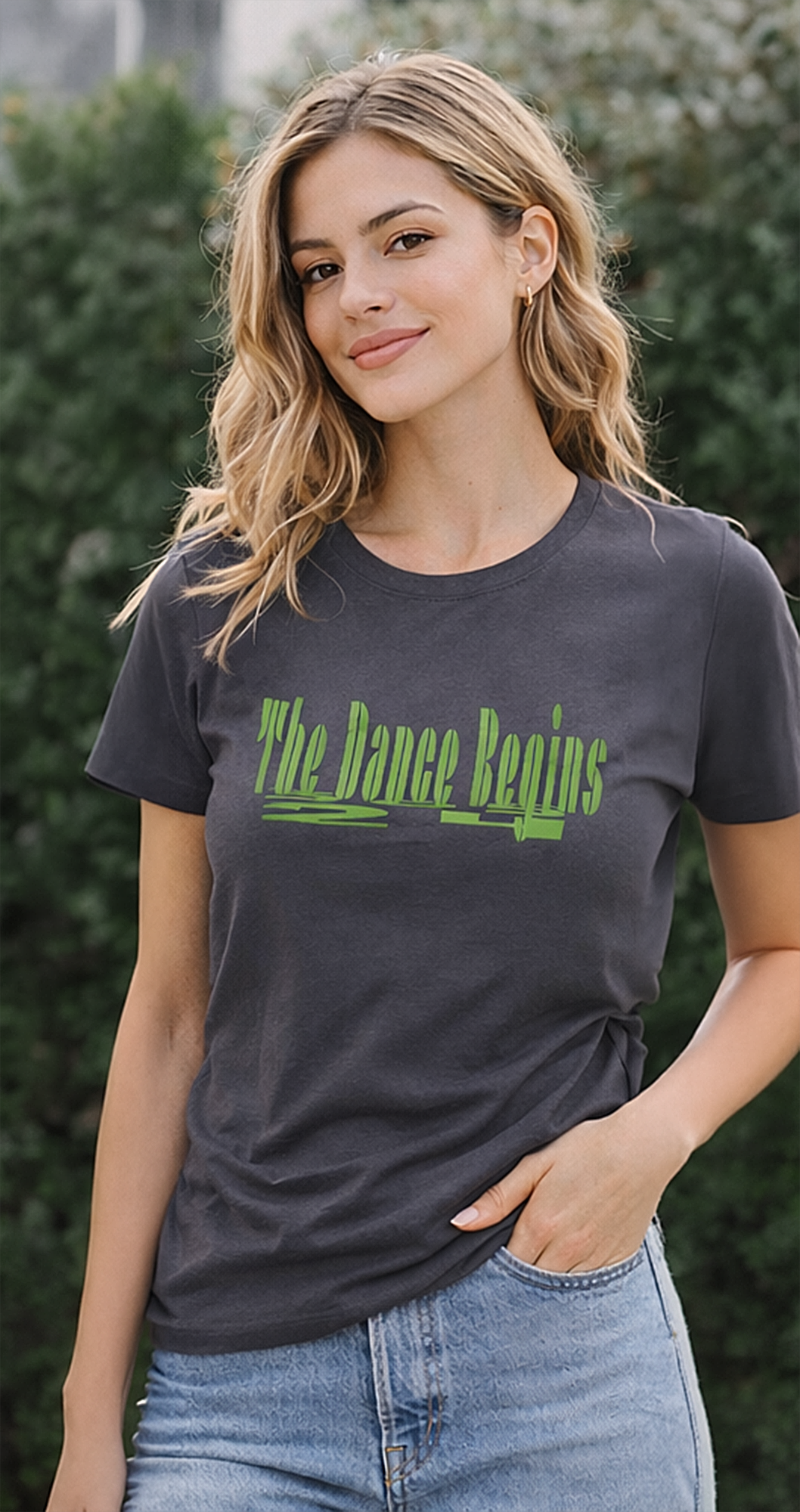 Dark gray t-shirt with the text "The Dance Begins" in green. The shirt is a crew neck style and is paired with blue denim jeans. The image is taken outdoors with a green leafy background.