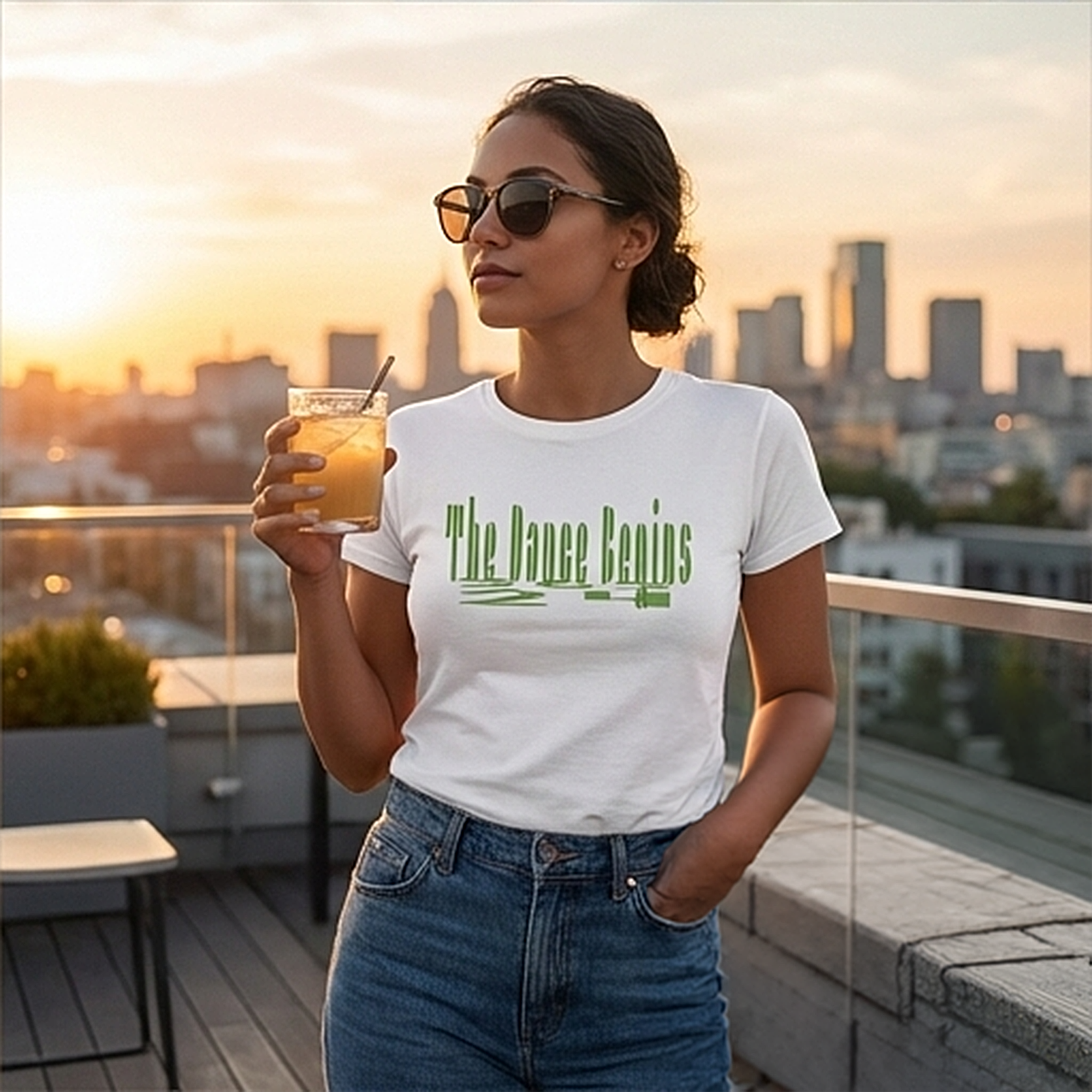 White t-shirt with the text "The Dance Begins" in green, paired with blue jeans. The image shows a person wearing sunglasses and holding a drink, set against a city skyline at sunset.