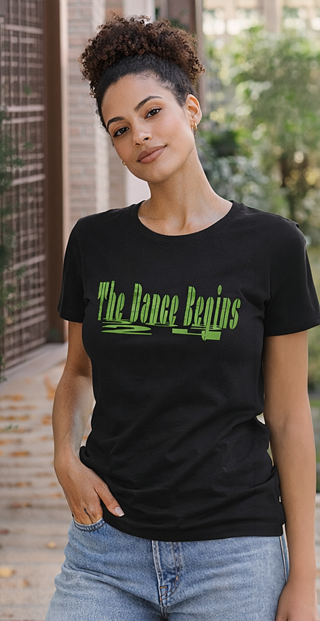 Black t-shirt with the text "The Dance Begins" in green. The shirt is worn by a person with curly dark hair and blue jeans. The person is standing outside near a brick wall.
