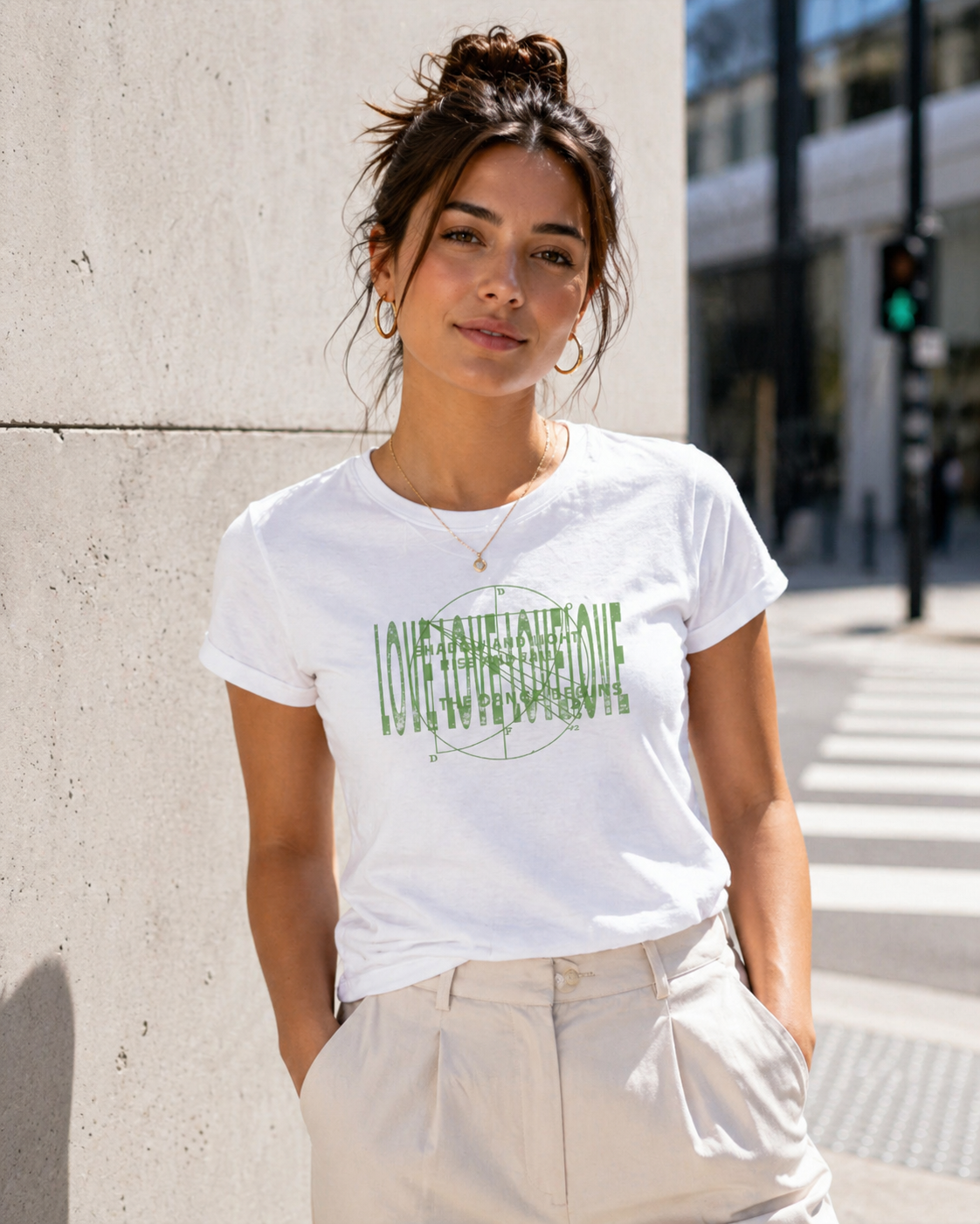Crew neck white t-shirt with a green texts and circular overlaid graphic design. A woman walking on an urban street wearing beige pants with one hand in her pocket.