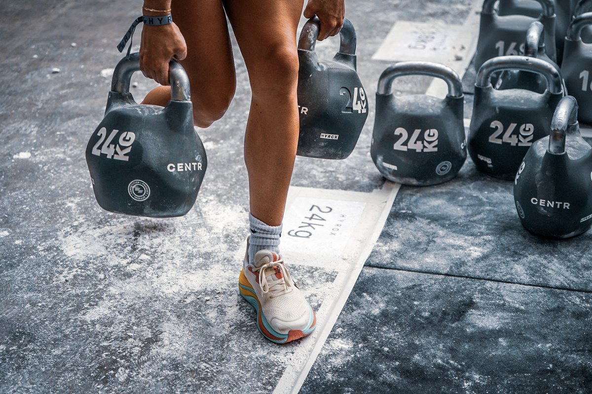 A person holding two kettlebells in each hand during a workout at the gym, with more kettlebells on the floor around them.