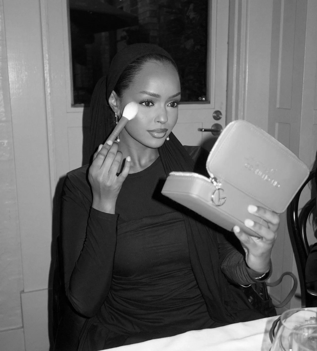 Woman with dark hair and earrings applying makeup while looking into a mirror, holding a makeup palette, in a room with a door and window in the background