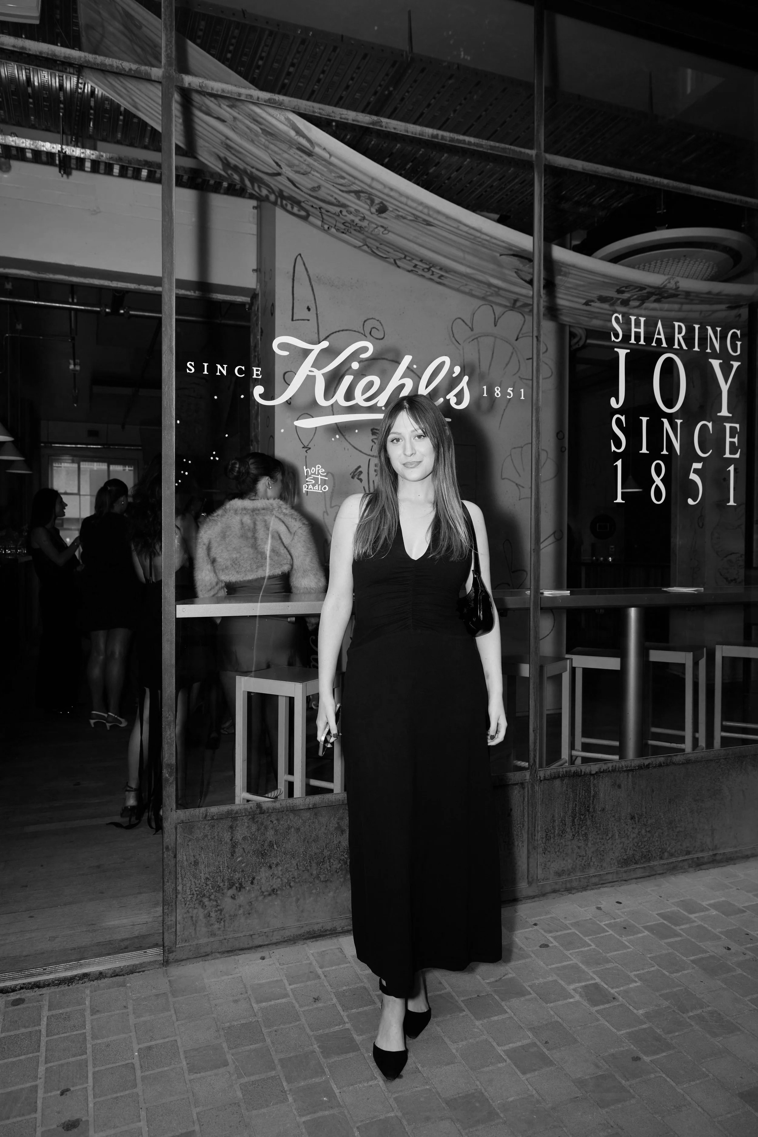 A woman in a black dress and high heels standing in front of a mirror, with a background showing people inside a store and a sign that reads 'Kiehl's Since 1851' and 'Sharing Joy Since 1851'.