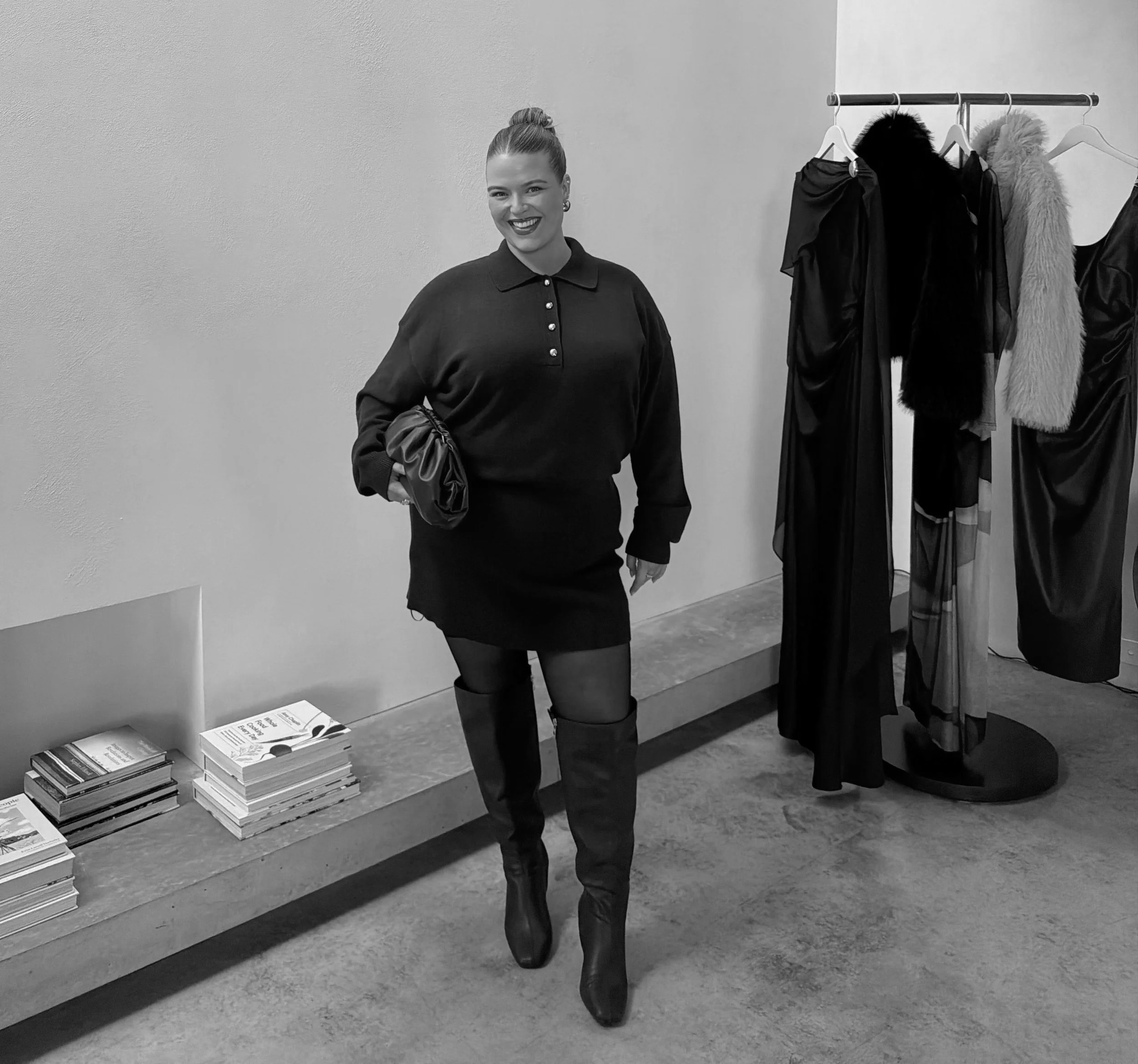 A woman in a dark dress and over-the-knee boots standing next to a clothing rack with black and fur-lined dresses or coats in a retail store.