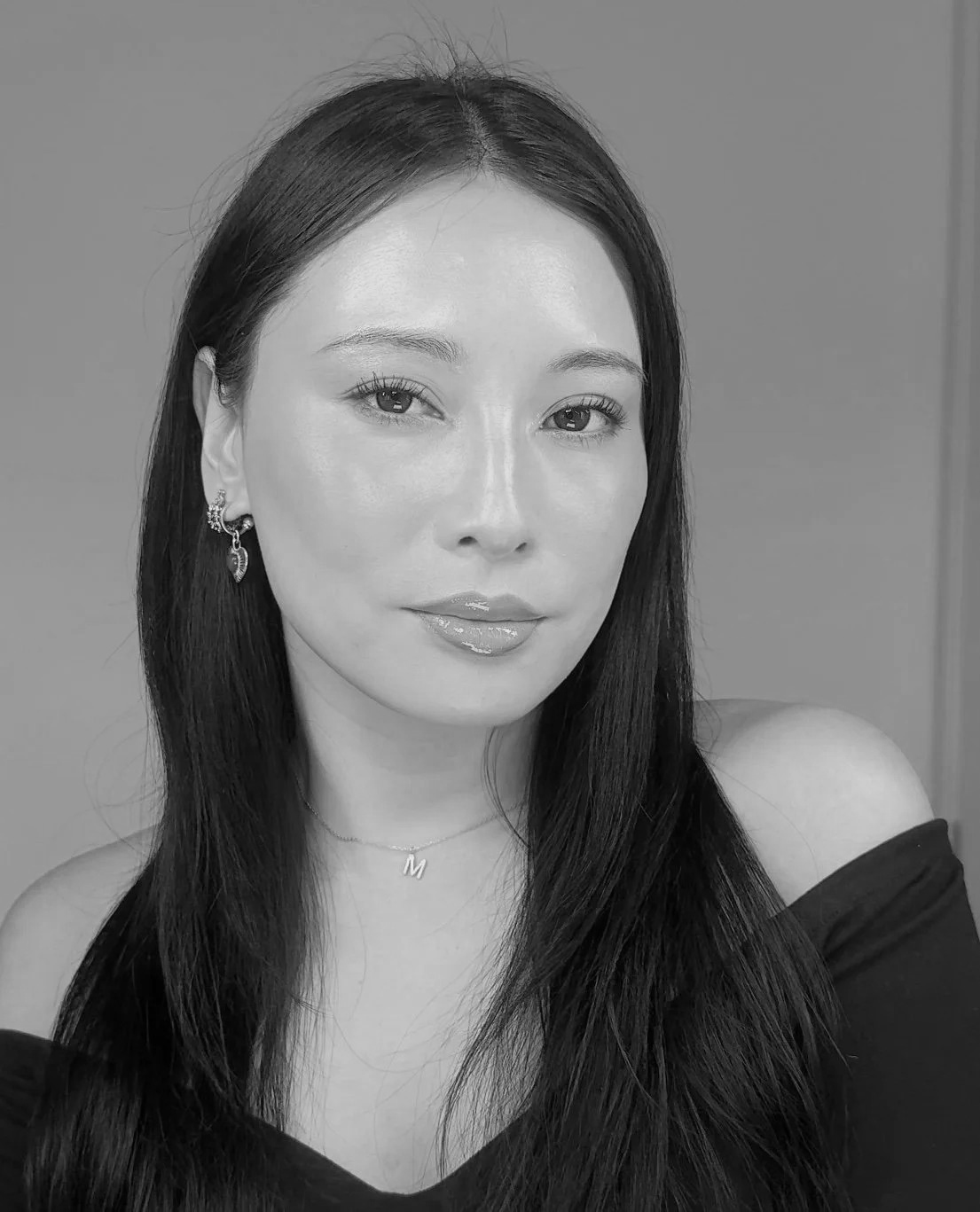 Black and white portrait of a woman with long dark hair, wearing earrings, a necklace with the letter 'M', and an off-the-shoulder top, looking at the camera.