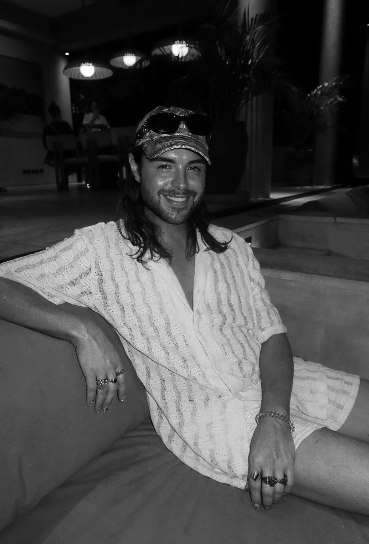 A man with long hair and a beard, wearing sunglasses on a cap and a light-colored, striped shirt, sitting on a couch in a dimly lit indoor space, smiling at the camera.