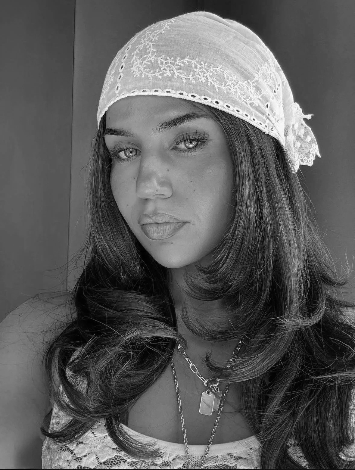 A woman with long wavy hair wearing a patterned headscarf and layered necklaces, posing for a portrait in black and white.