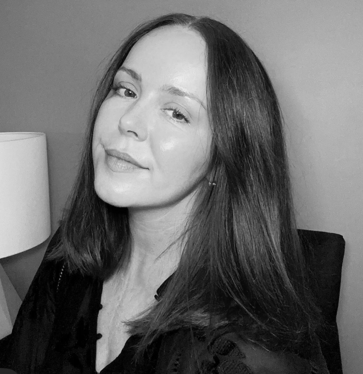 A woman with long dark hair, wearing a dark outfit, posing in front of a plain wall with a lamp visible in the background.