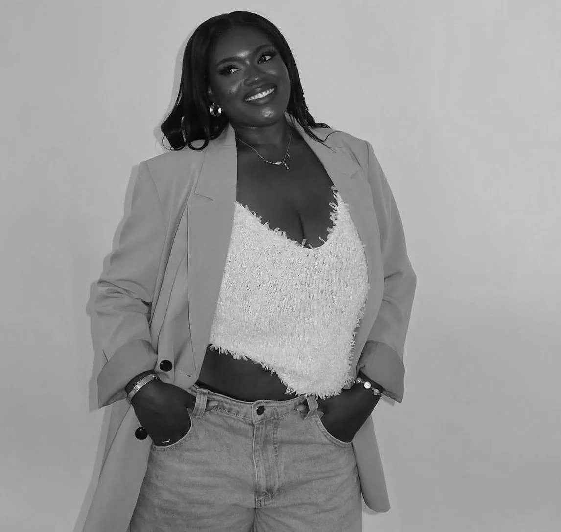 A smiling woman dressed in a blazer, fuzzy top, and jeans, standing against a plain background.