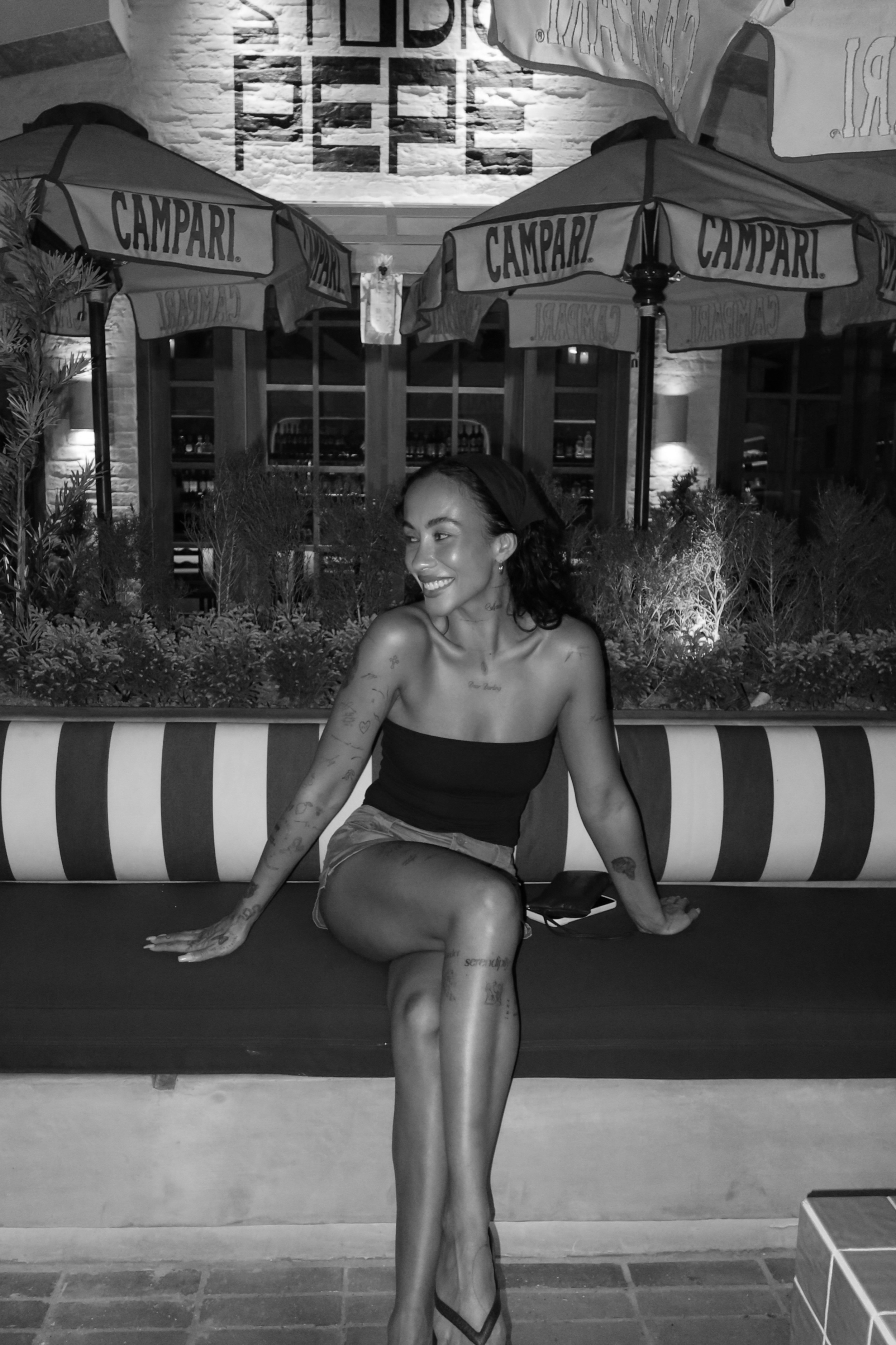 A woman with tattoos and a black headband sitting on a bench with one leg crossed over the other, smiling, under umbrellas with 'CAMPARI' logos, in an outdoor cafe at night.