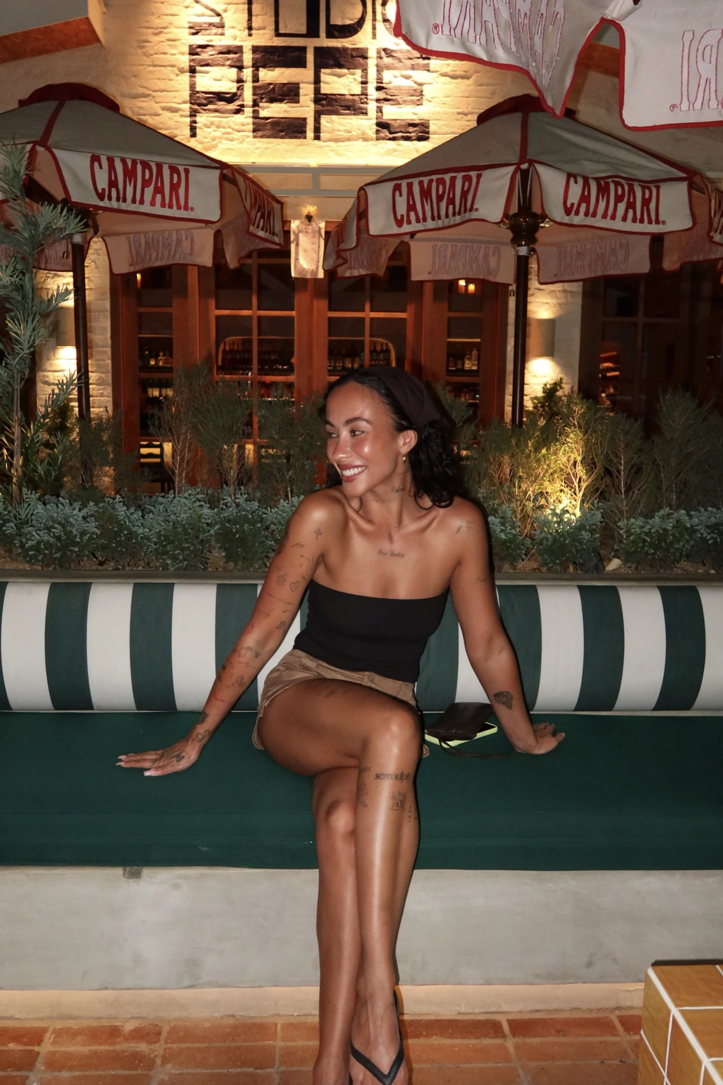 A woman with dark curly hair and multiple tattoos sitting on a green striped bench in an outdoor restaurant or patio area at night. She is wearing a black strapless top, beige shorts, and black flip-flops while smiling and looking to her right, with her left arm resting on her knee and her right hand on the bench. In the background, there are umbrellas with 'CAMPARI' branding, lush plants, a brick wall, warm lighting, and wooden wine racks.
