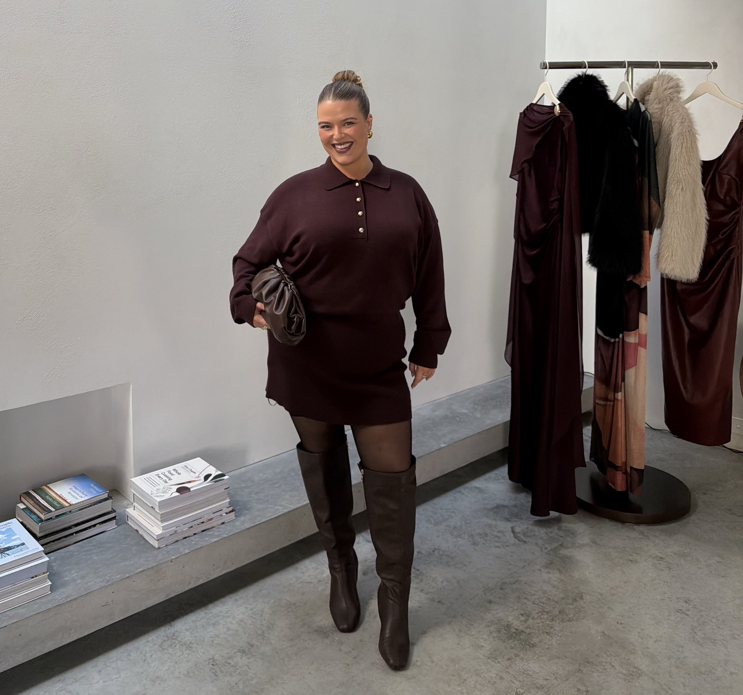A woman with a bun hairstyle wearing a dark burgundy dress with gold buttons, black tights, and knee-high black leather boots, standing next to a clothing rack with various dresses and coats, and a stack of books on the floor.