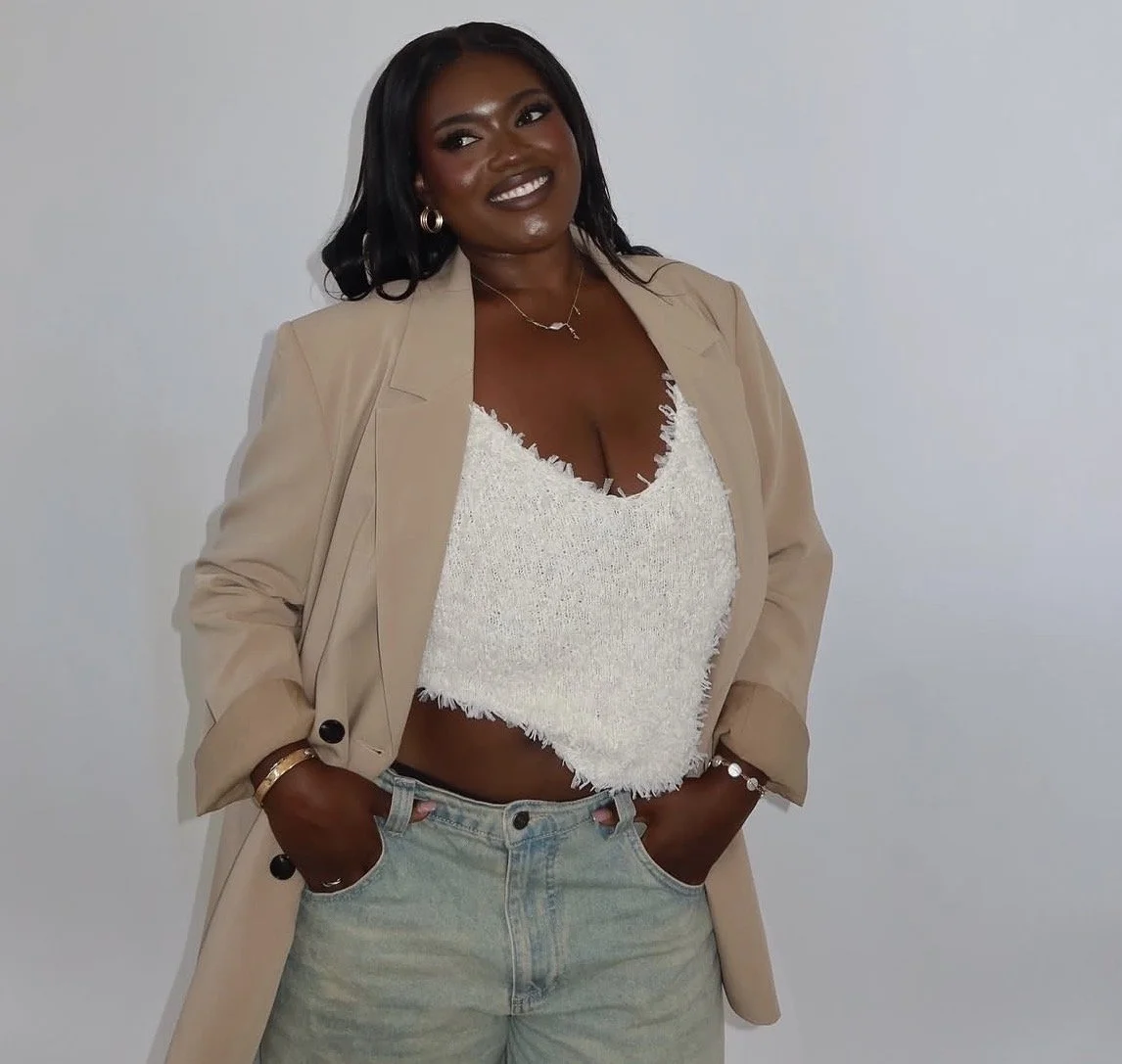 A woman with dark skin and black wavy hair standing against a plain white wall, wearing a beige blazer, a white textured crop top, light-wash jeans, gold jewelry, and smiling.
