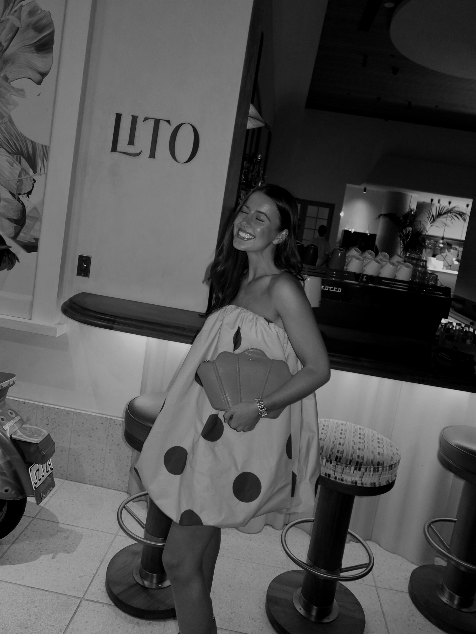 A woman smiling and posing for the camera in a café, holding a polka dot bag, standing next to a barstool, with a LUTO sign on the wall behind her, and the café interior visible in the background.