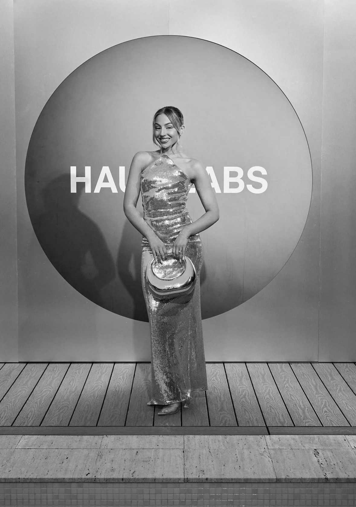 A woman in a shiny, sequined gown standing in front of a circular sign that reads 'HAVE AN AB.' She is holding a matching shiny clutch and smiling, with a wooden floor and wall in the background.