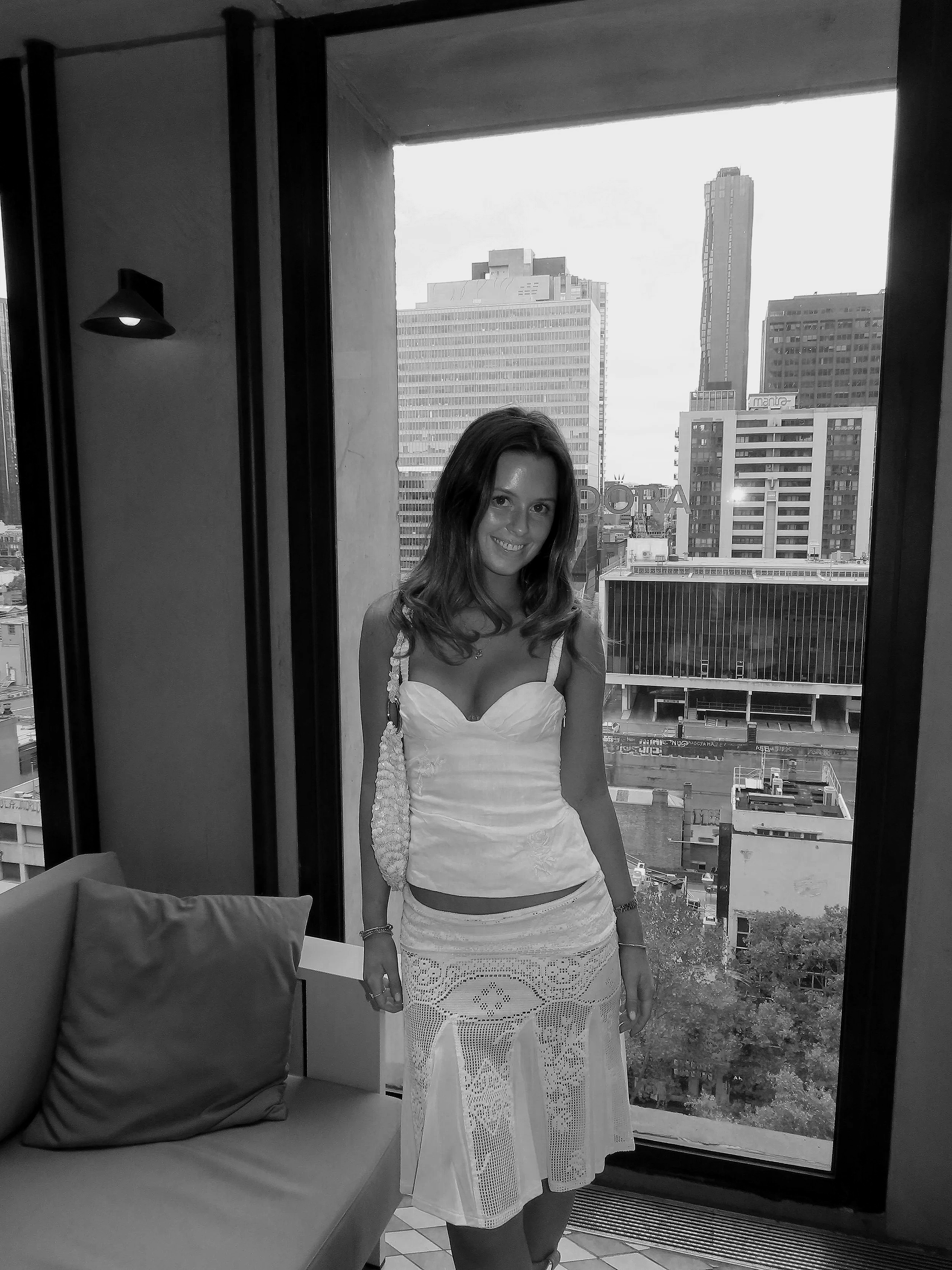 A woman standing near a large window in a high-rise building, smiling, with a cityscape of tall buildings and skyscrapers in the background.