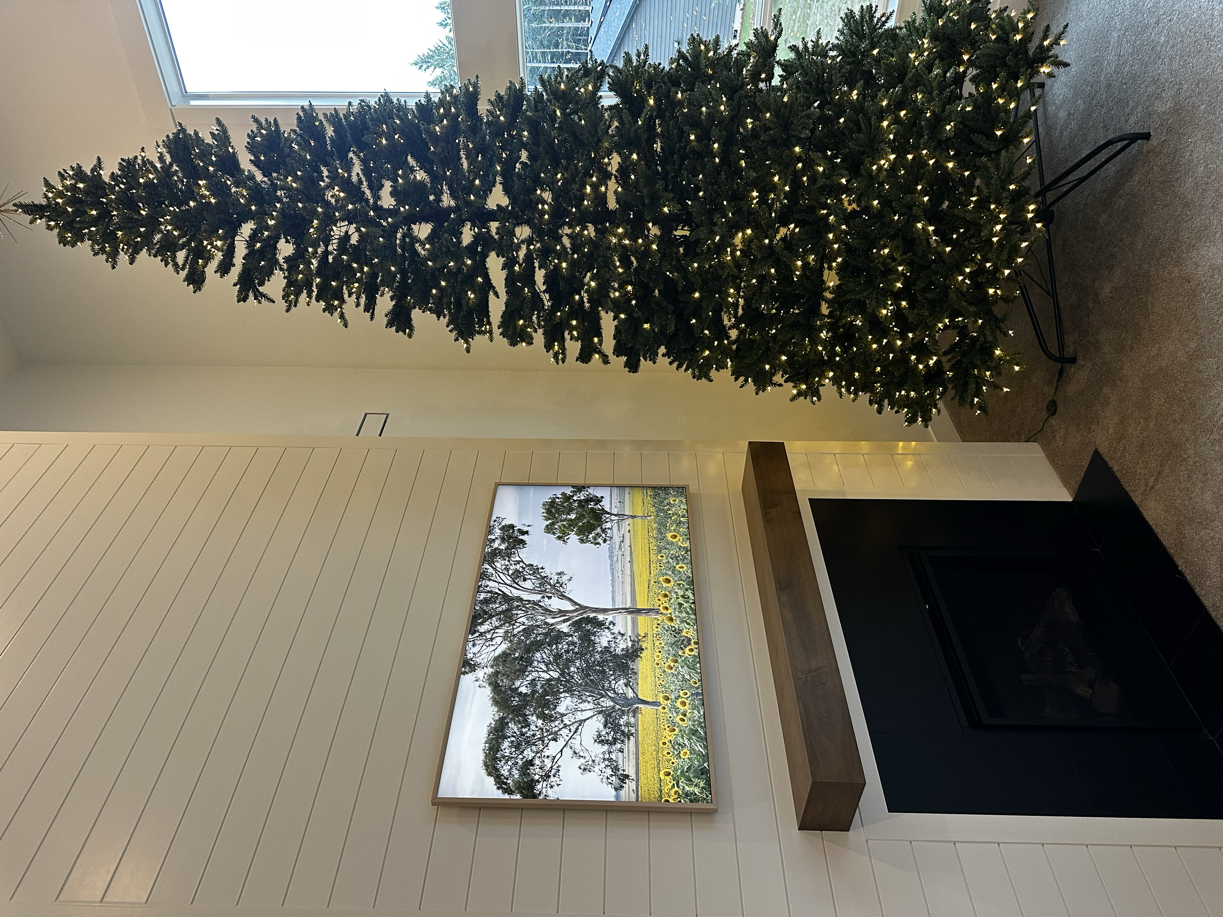 A decorated Christmas tree with lights stands next to a white wall with a window above, a fireplace, and a landscape painting featuring trees and a field of sunflowers.
