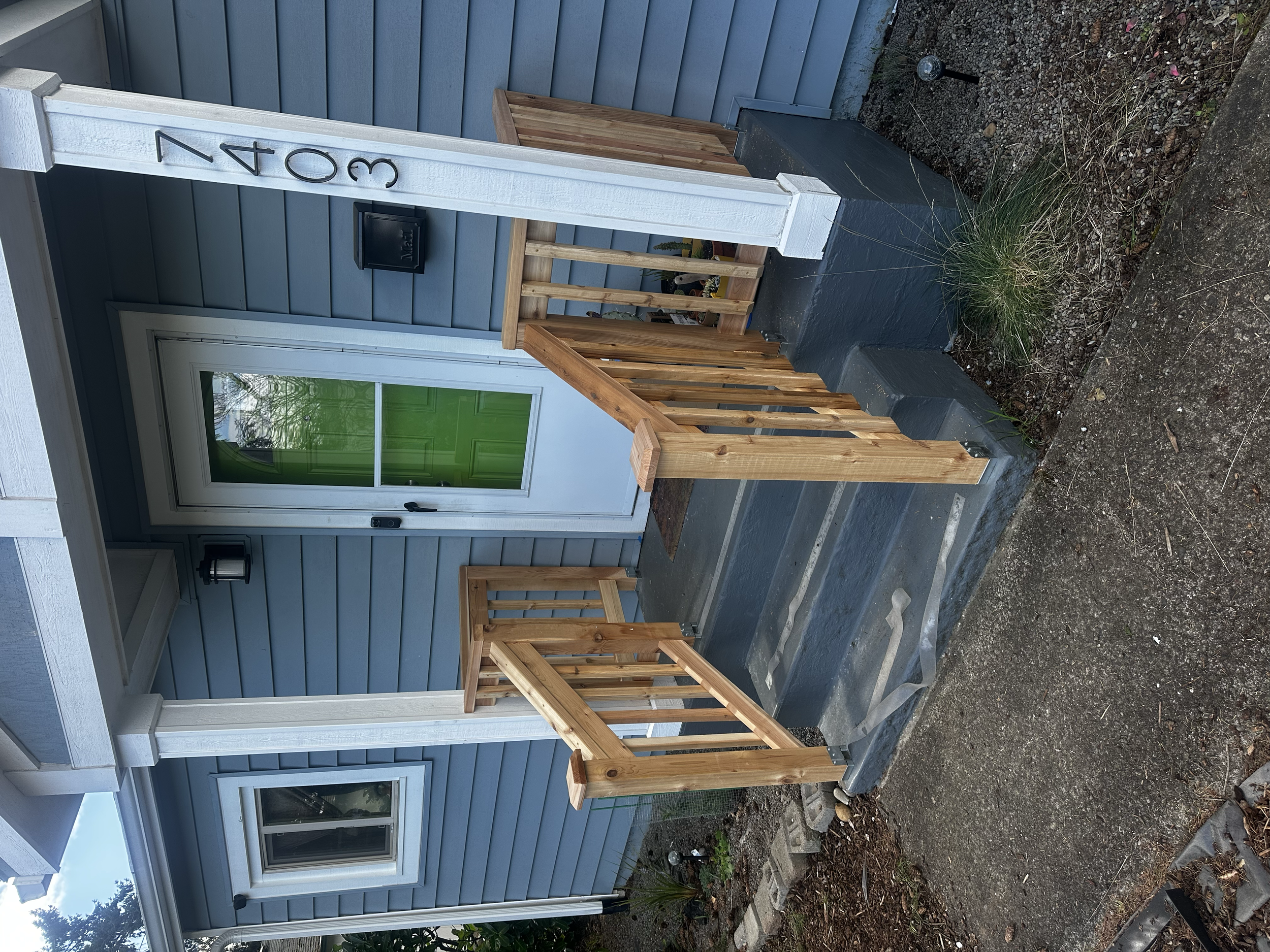 Front porch with a blue house, a white door with a green window, and a small staircase with wooden handrails leading to the entrance.
