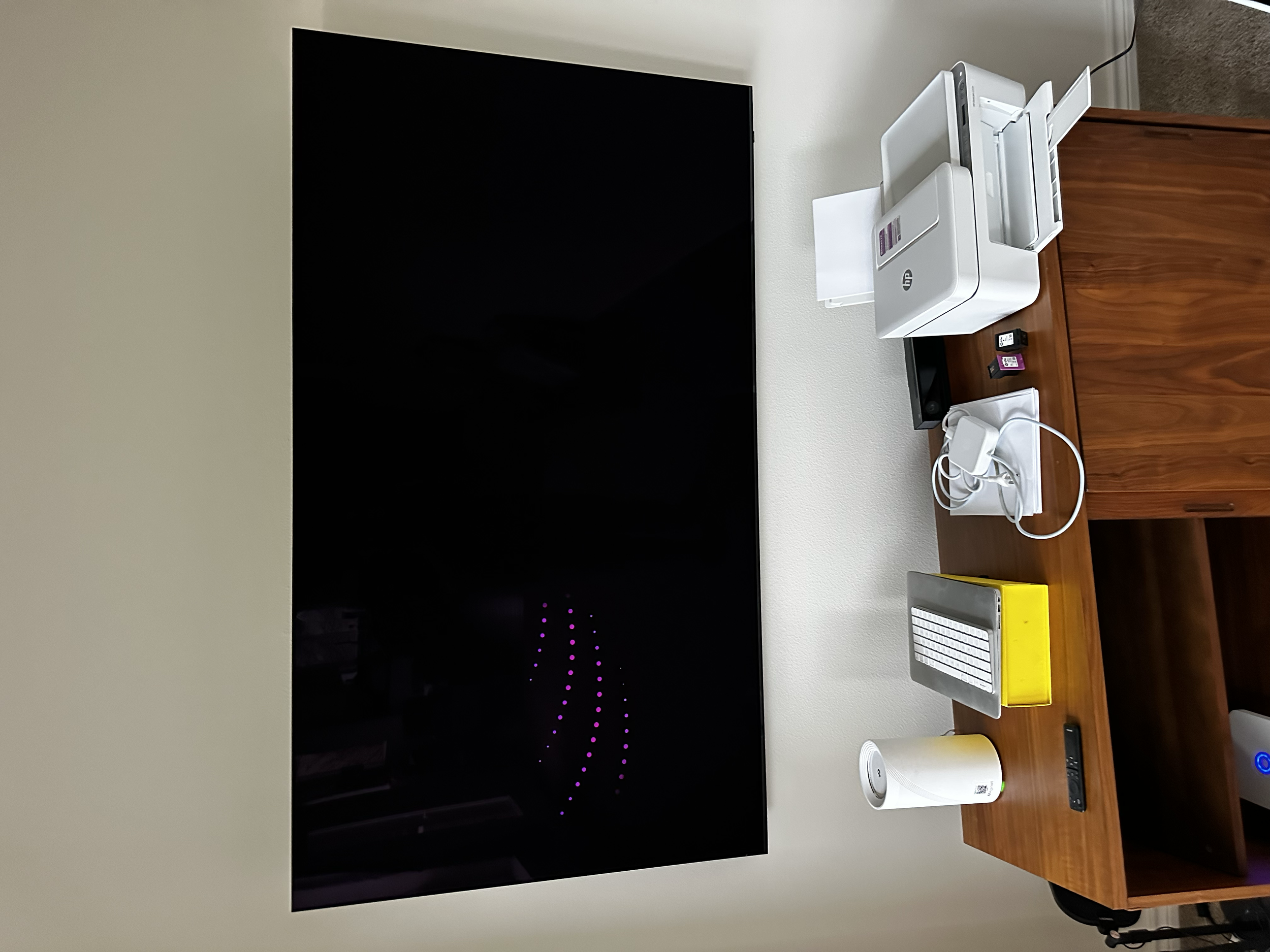 Desk with a large flat-screen TV, a white HP printer, various small electronic devices, a yellow box, a white cup, and a wireless keyboard on a wooden desk.