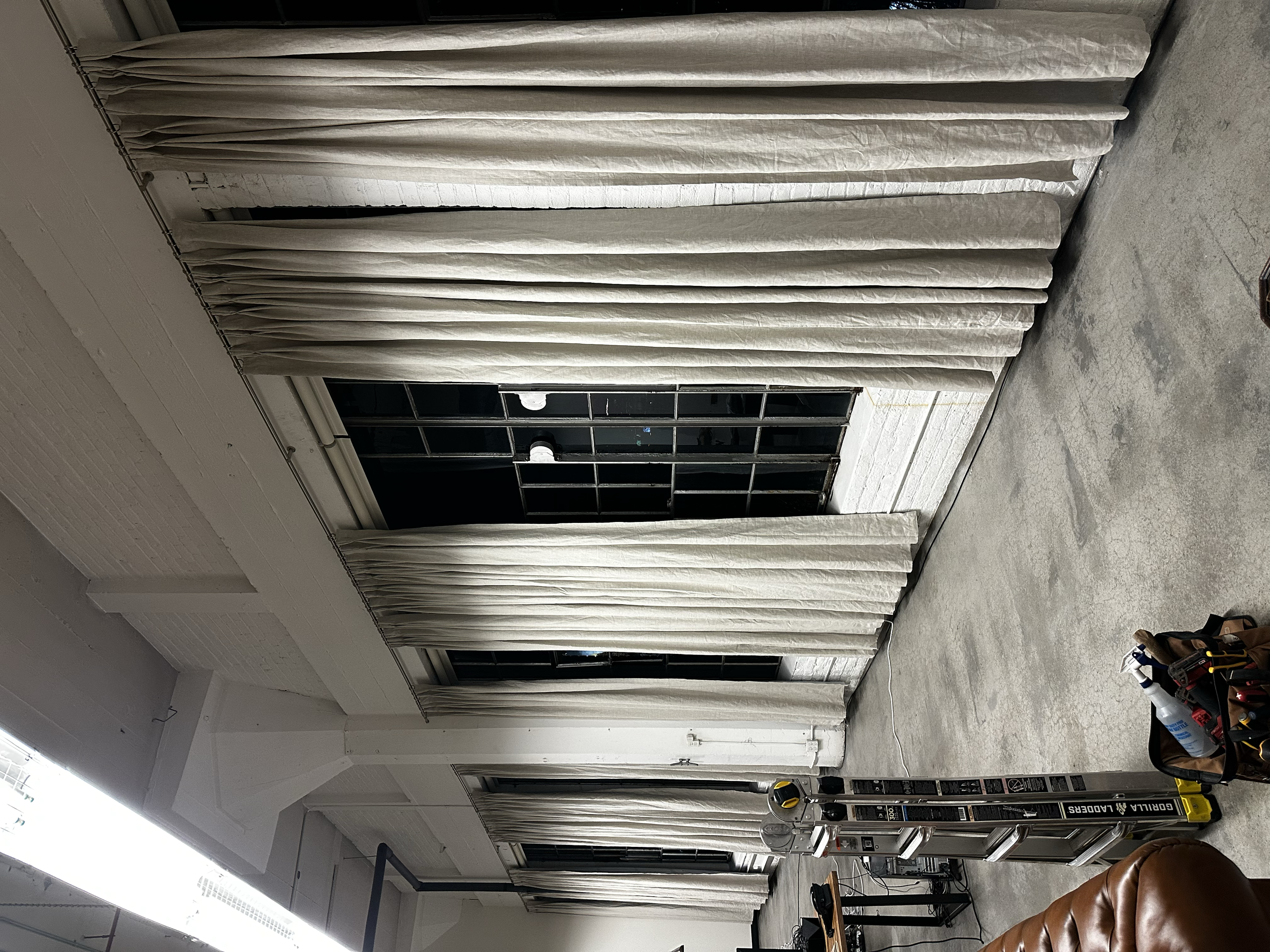Empty industrial space with beige curtains covering large windows, concrete floor, ladder, and tools on the right side.