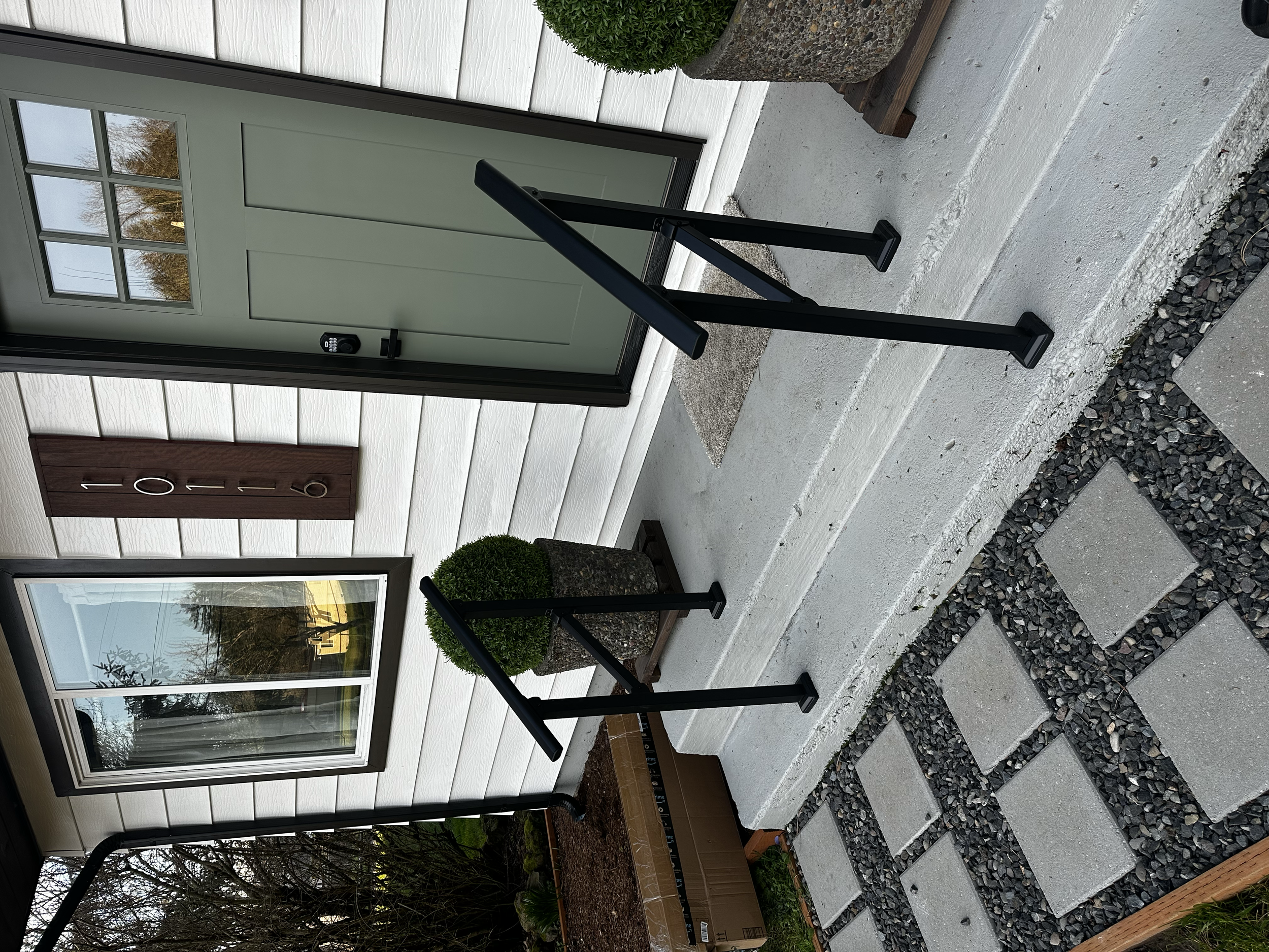 Front porch area with a green door, window, and house number 1016. A metal handrail leading to the door and a potted plant with a trimmed bush are visible. The ground has light-colored stairs and a walkway with grey pavers bordered by small black sto
