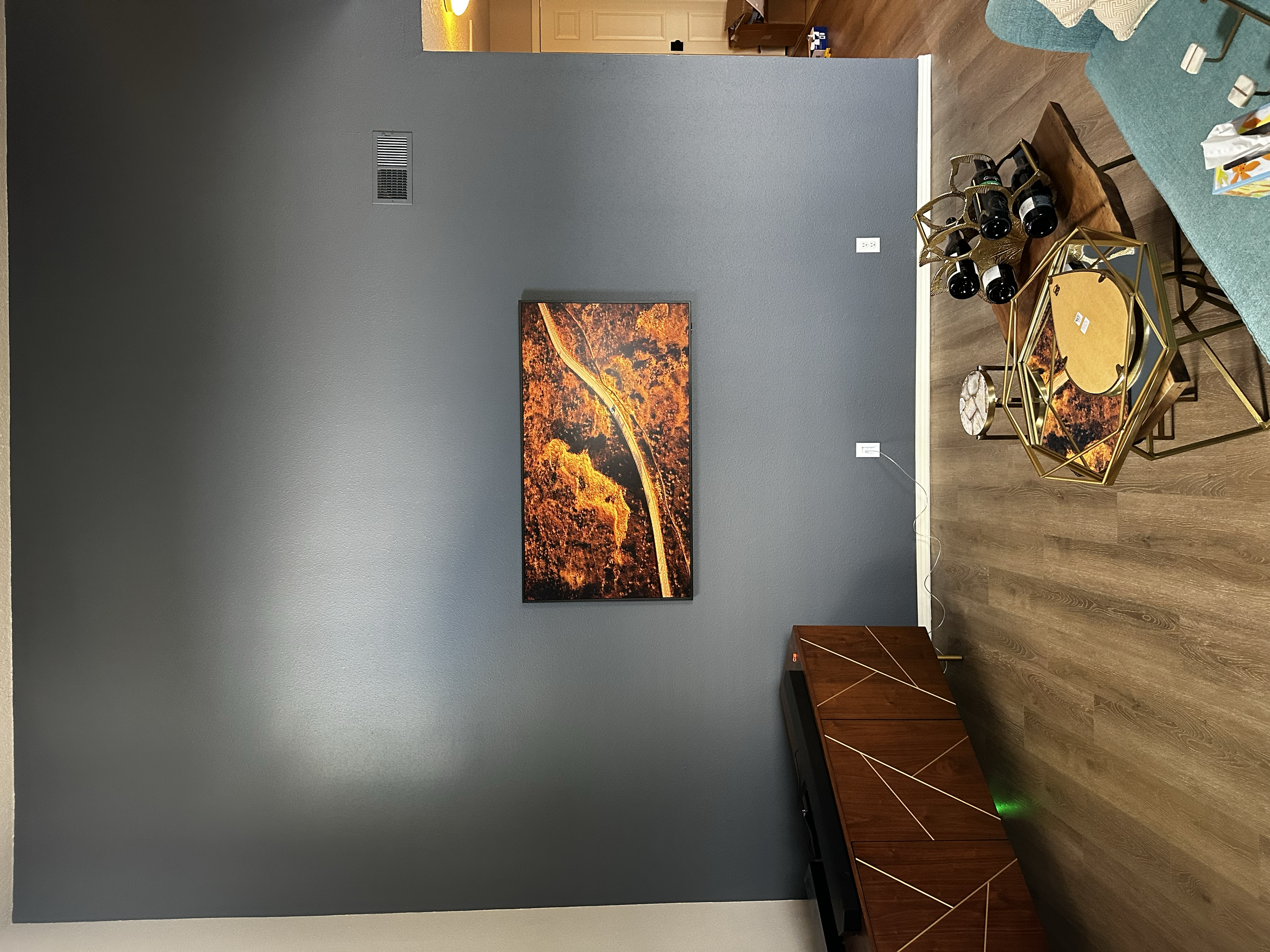 Living room with a dark gray wall, a framed aerial view of a winding road through an orange fall forest, a gold geometric side table layered with mirrors, bottles, and decorative items, and a wooden cabinet below the wall art.