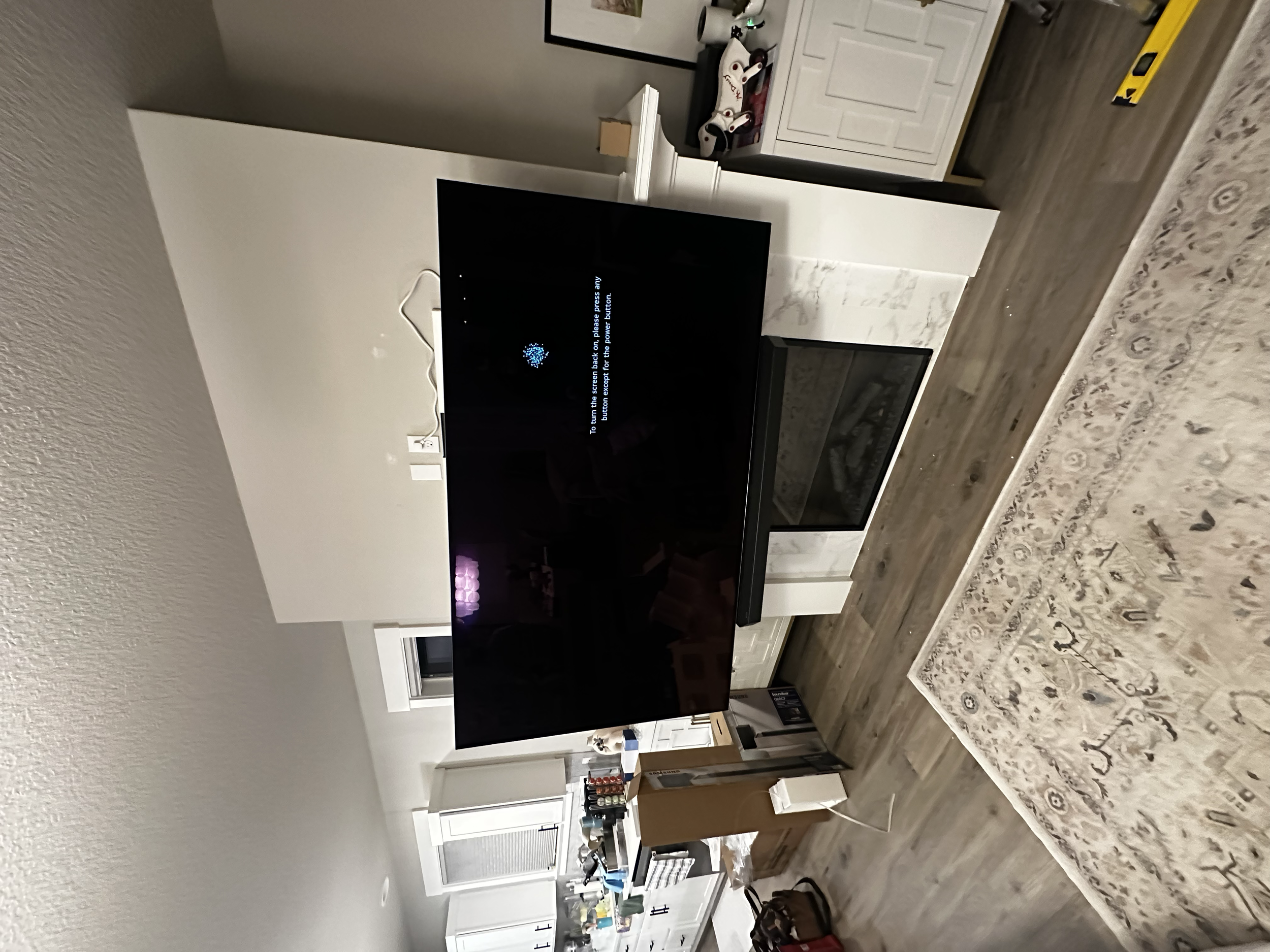 Living room with a large flat-screen TV on a white fireplace mantel, showing a blue loading icon and message. To the right of the TV is a white cabinet with golf shoes and other items on top. There is a patterned area rug on wooden floor and a window