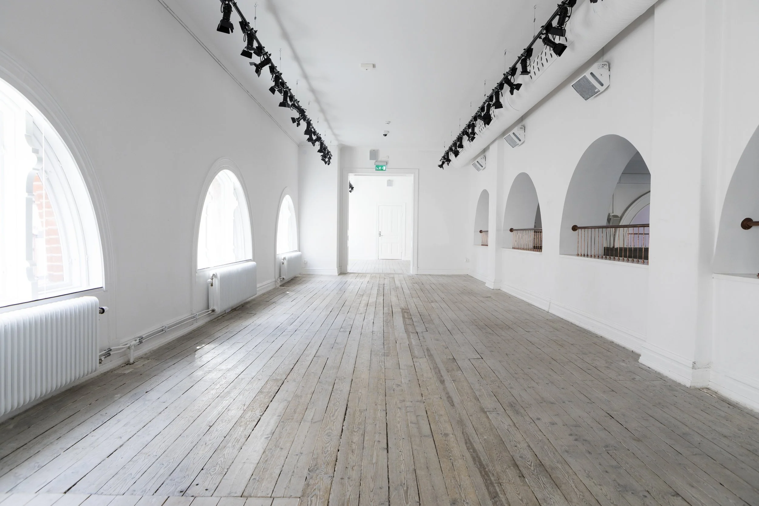 Ett tomt rum med vita väggar, stora fönster, ljus trägolv och takbelysning på spår.