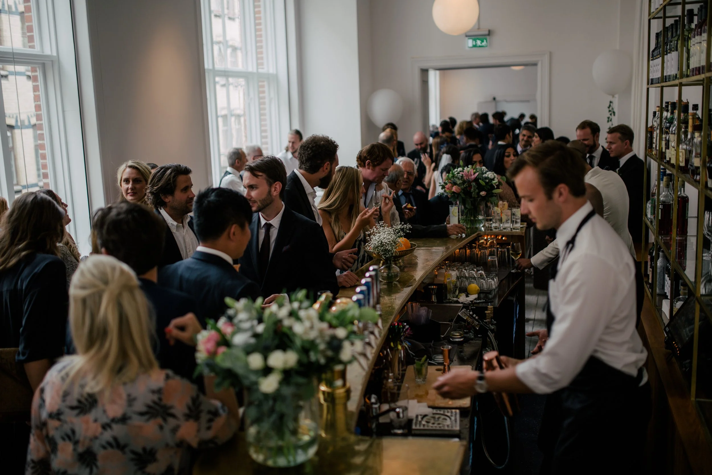 Många människor i formella kläder samlas i ett rum, står vid en bar med blommarrangemang och serveringspersonal.