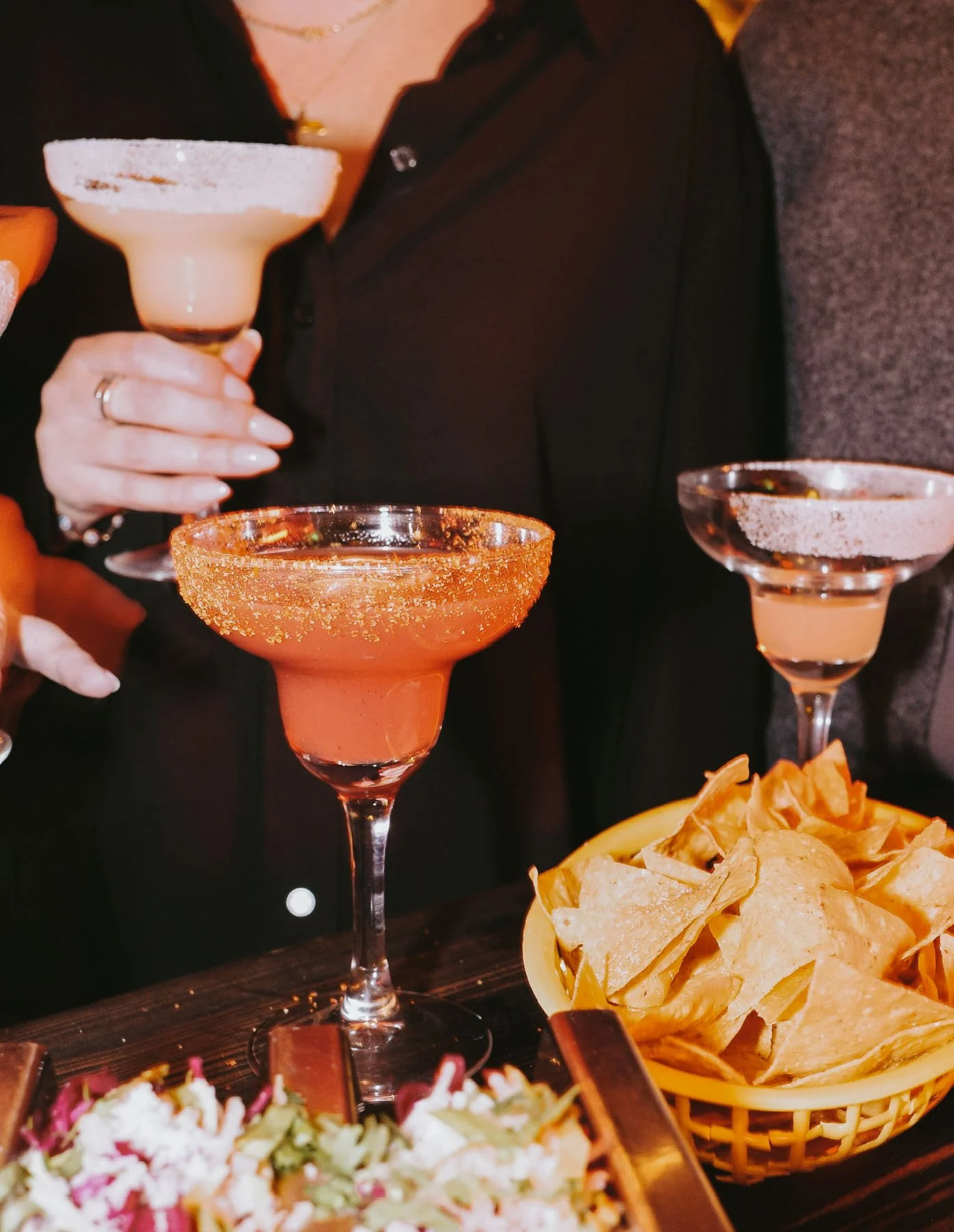 Person håller tre drinkar i cocktailglas, en på varje hand, med snacks på bordet.