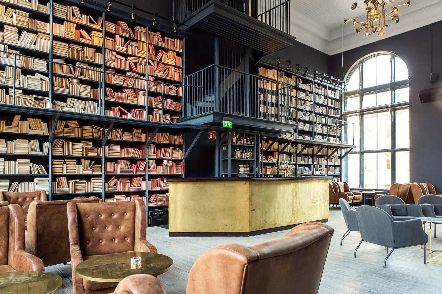 Inomhusbar i ett bibliotek med höga bokhyllor fyllda med böcker, komfortabla fåtöljer, runda bord, stora fönster och modern belysning.