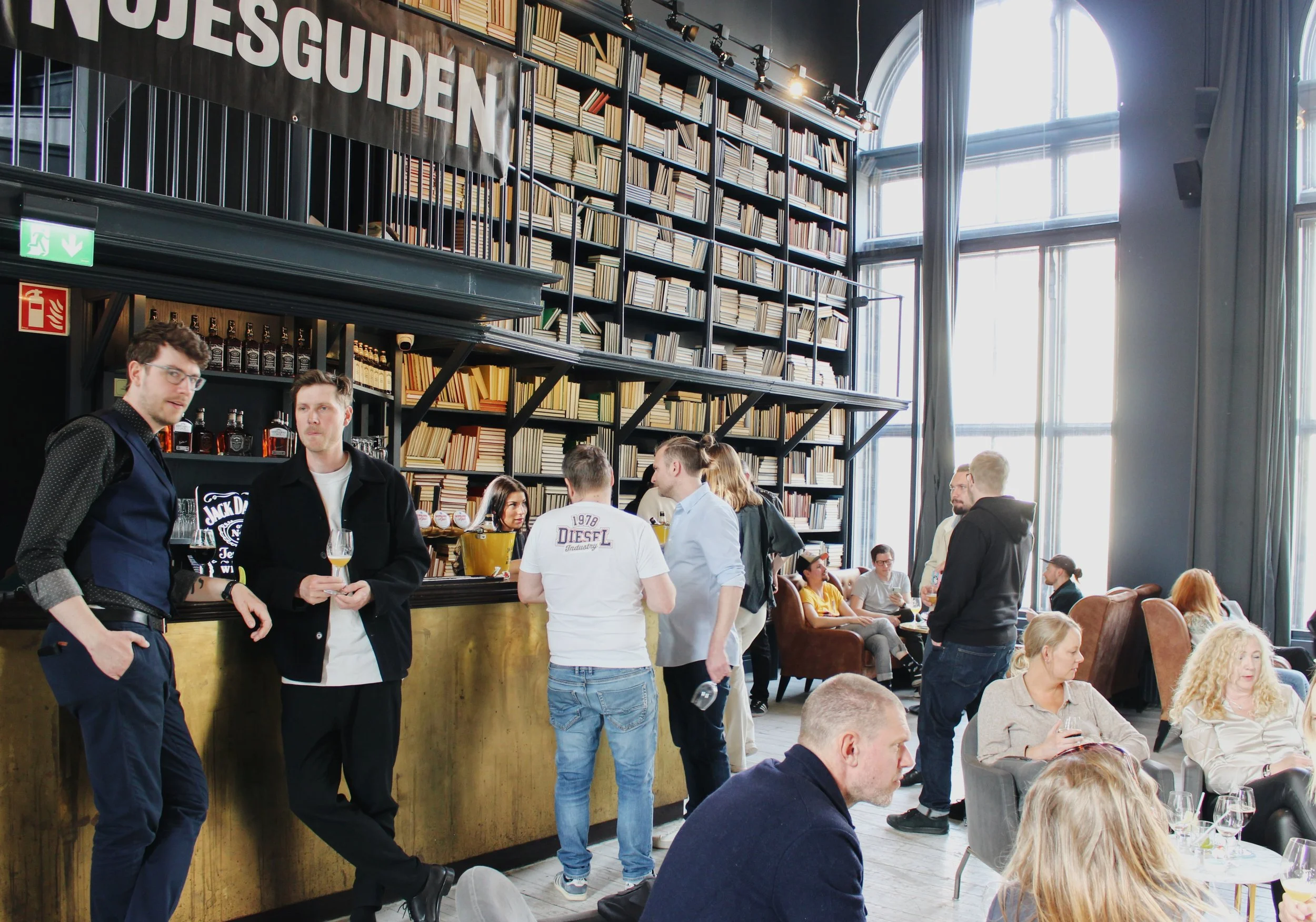 Människor som umgås och dricker i ett café med stora fönster och väggar fyllda med böcker, baren är förgylld, och det finns en stor banner med texten 'FÖRENINGEN'.