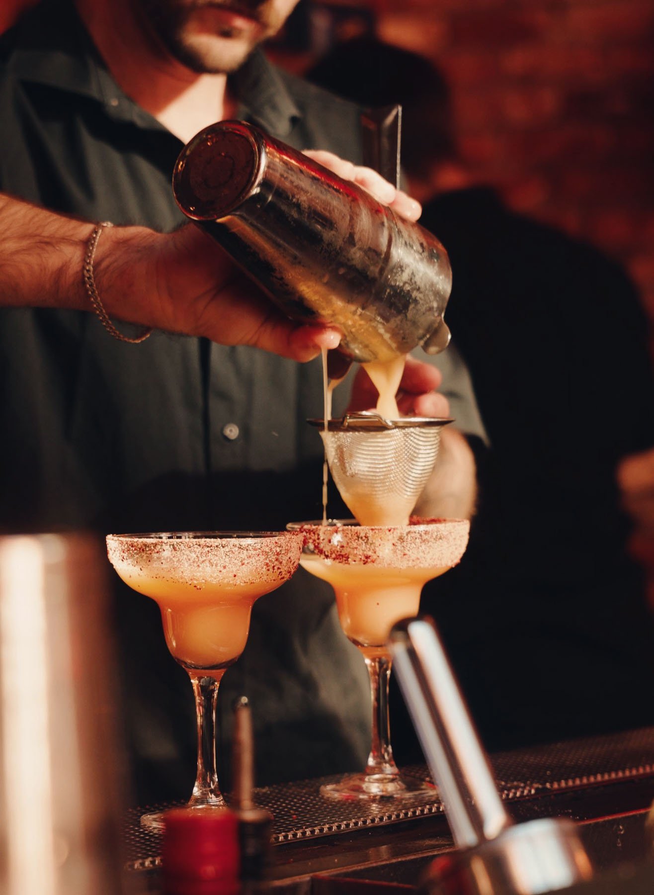 En bartender häller en drink genom en sil i två margarita-glass som är dekorerade med röda kryddor på kanten, i en bar med dimmig och varm belysning.