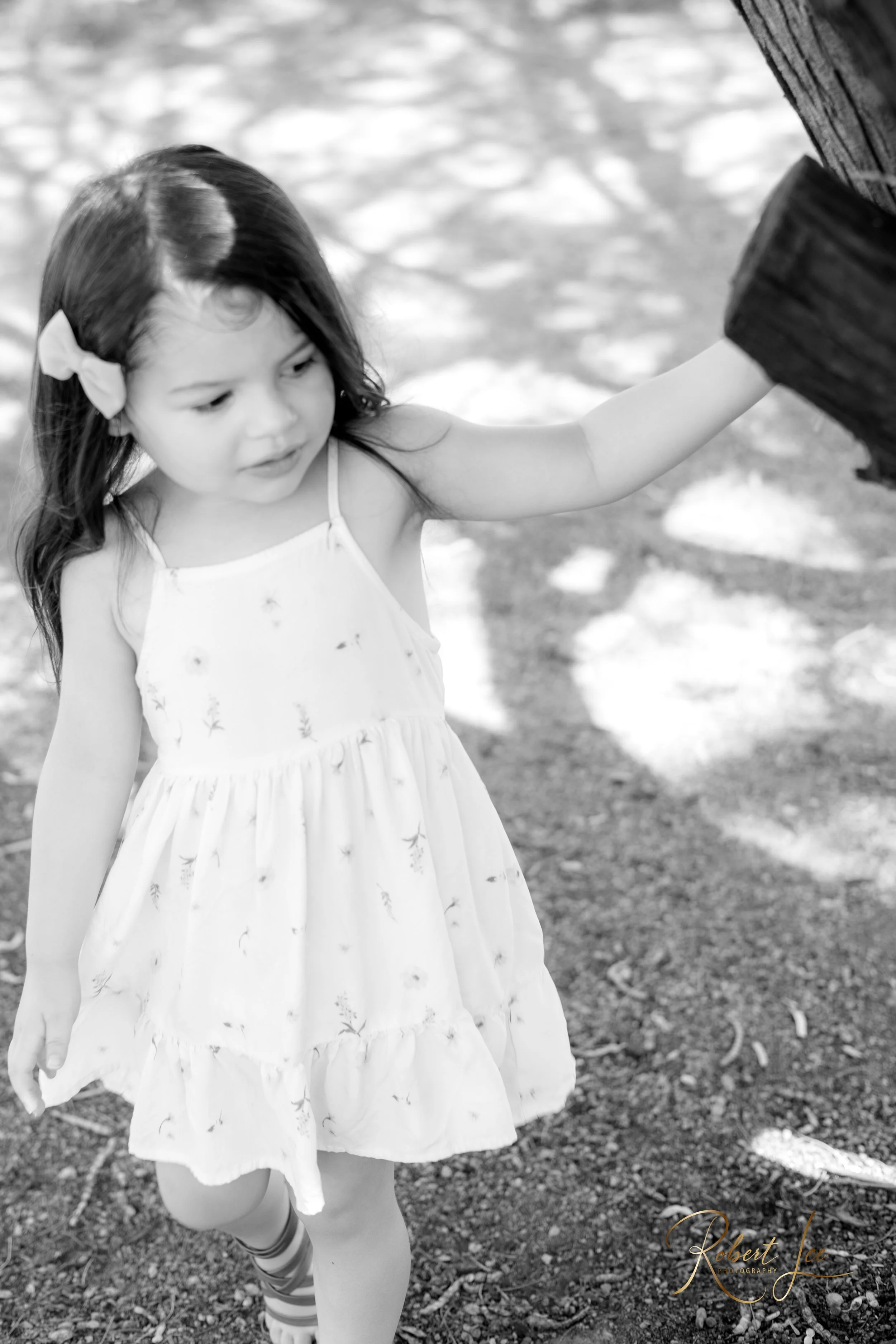 A young girl in a white dress with floral pattern and a bow in her hair stands outdoors, touching a tree with her right hand, on a sunny day. Tucson Portrait photographer. Robert lee Photography.
