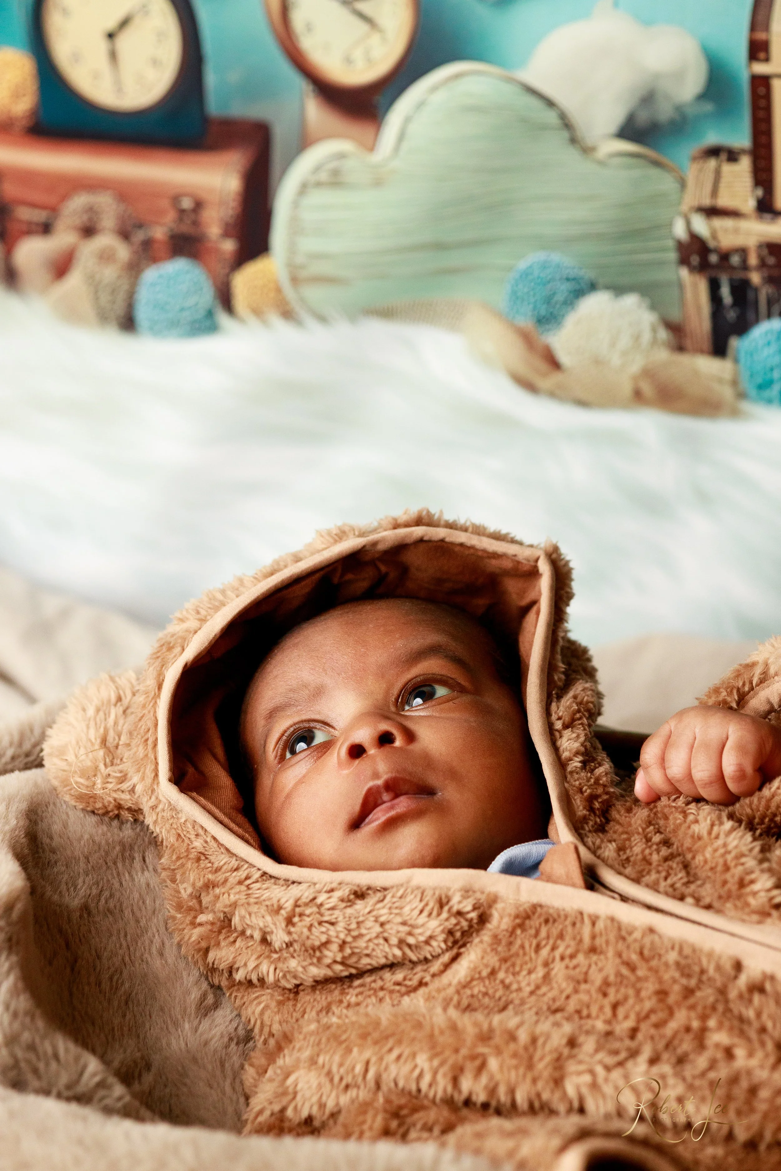 A baby dressed in a teddy bear costume lying in bed with a whimsical, colorful backdrop featuring clouds, clocks, and various plush toys.