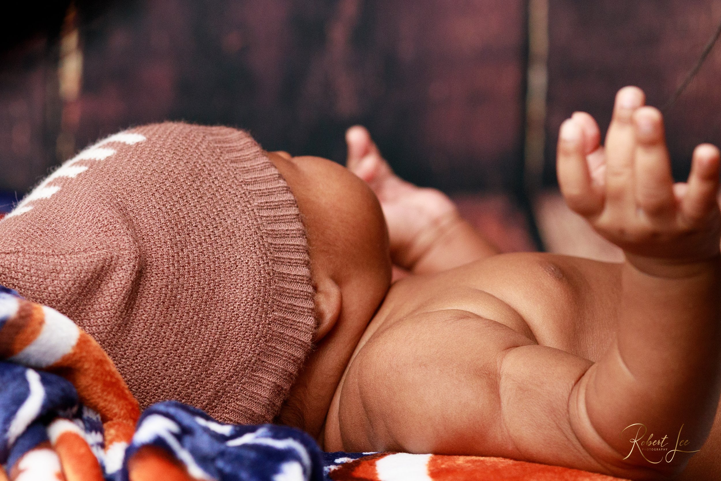 Close-up of a sleeping baby wearing a brown knit cap, lying on a colorful blanket with a dark background.