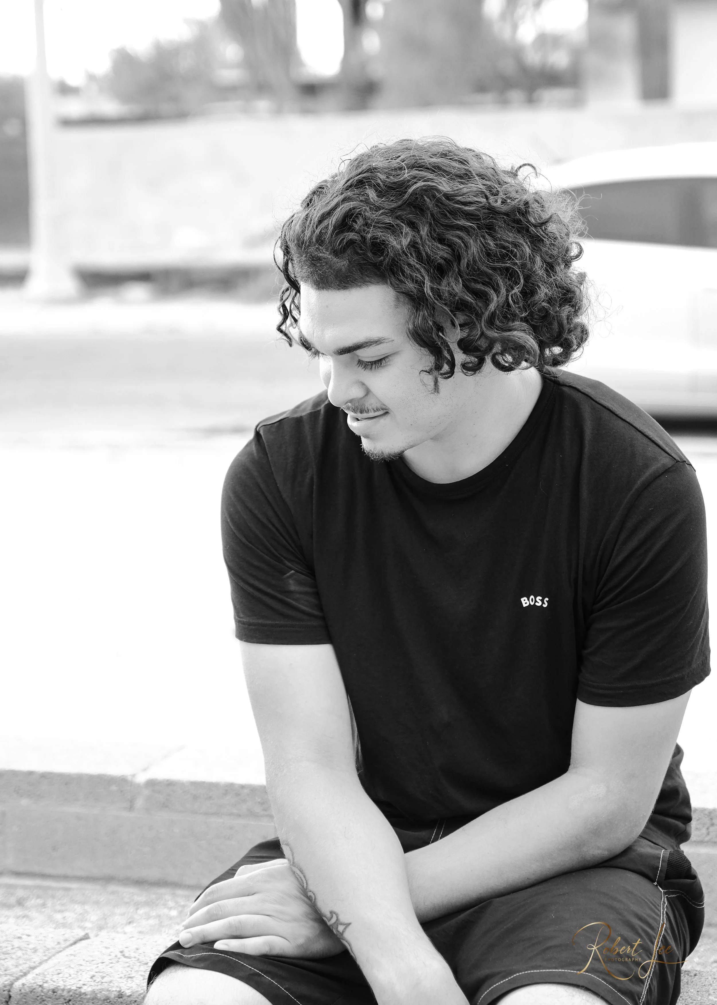 A young man with curly hair and tattoos on his arm sitting outdoors, looking down with a gentle expression, wearing a black T-shirt with the word 'BOSS' on it.