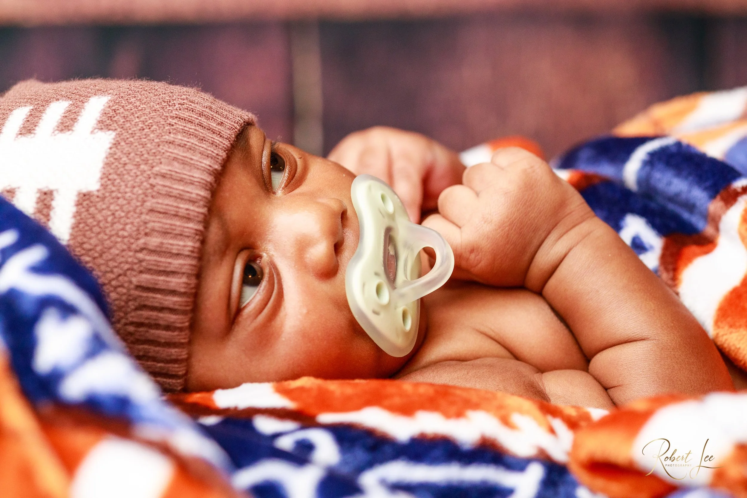 Close-up of a baby with brown skin lying on a blanket, wearing a pink knit hat, and using a pacifier.